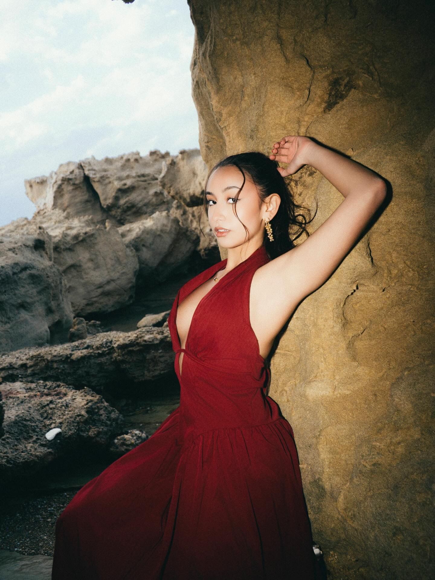 Woman in red halter dress poses against rocky coastal cliffs, gold earrings catching light in an elegant outdoor portrait.