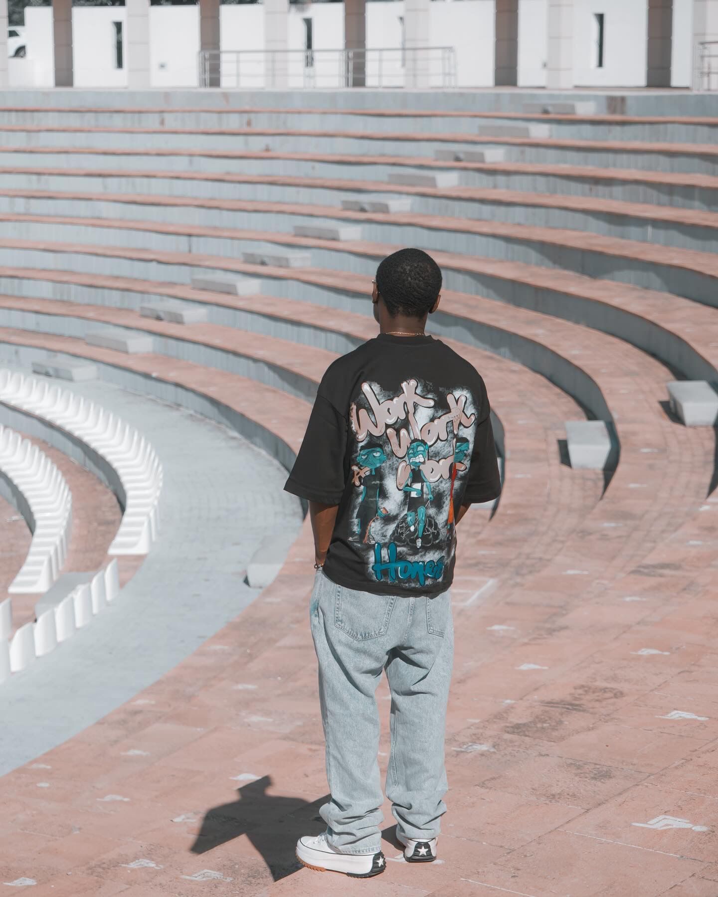 Man in black graphic tee and jeans stands in empty outdoor amphitheater, viewed from behind.