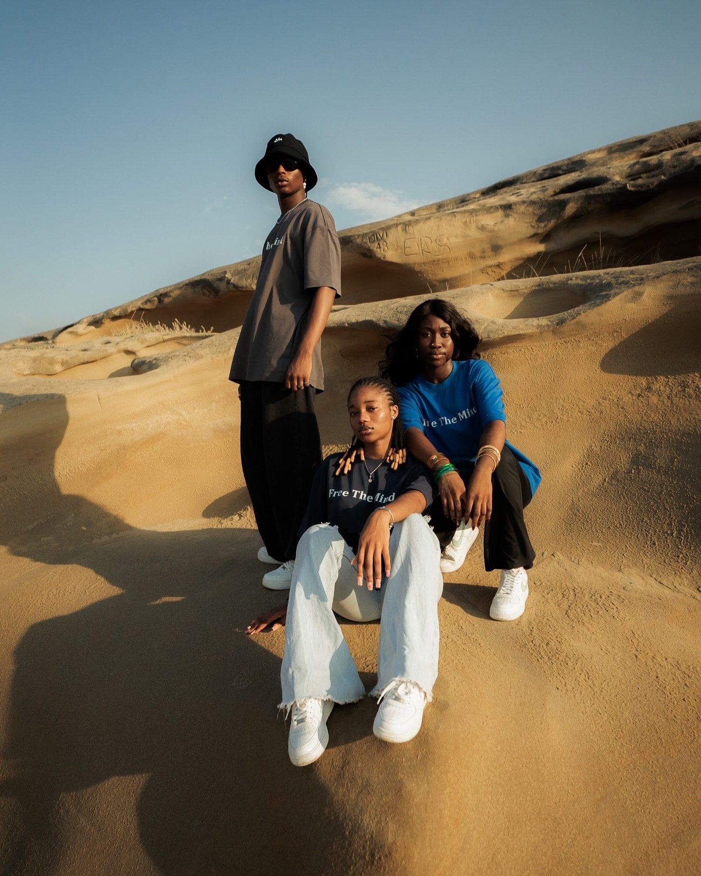 Three friends pose on sandy desert dunes, two standing and one seated, wearing casual streetwear in warm golden light.