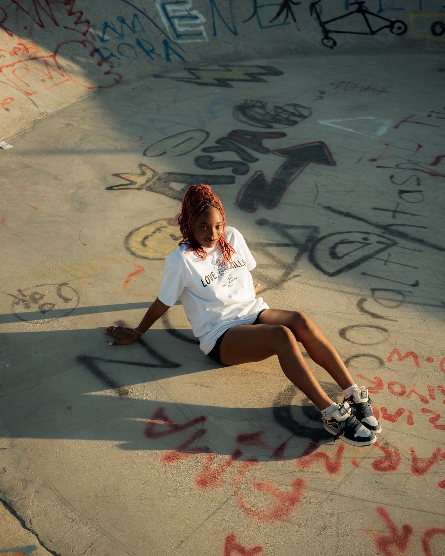 Young woman with red braids sits relaxed at a graffiti-covered skate park, wearing a white tee and sneakers.