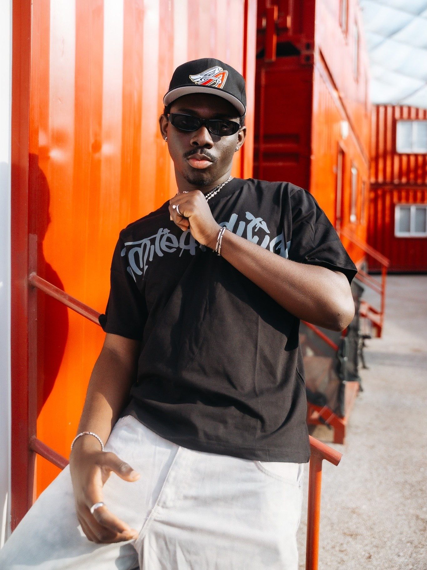 Young man in black graphic tee, white pants, cap and sunglasses posing against vibrant red wall outdoors.