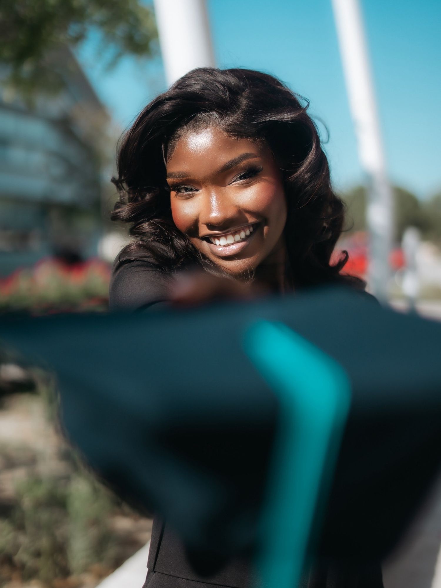 Smiling young Black woman holding graduation cap outdoors on a sunny day.