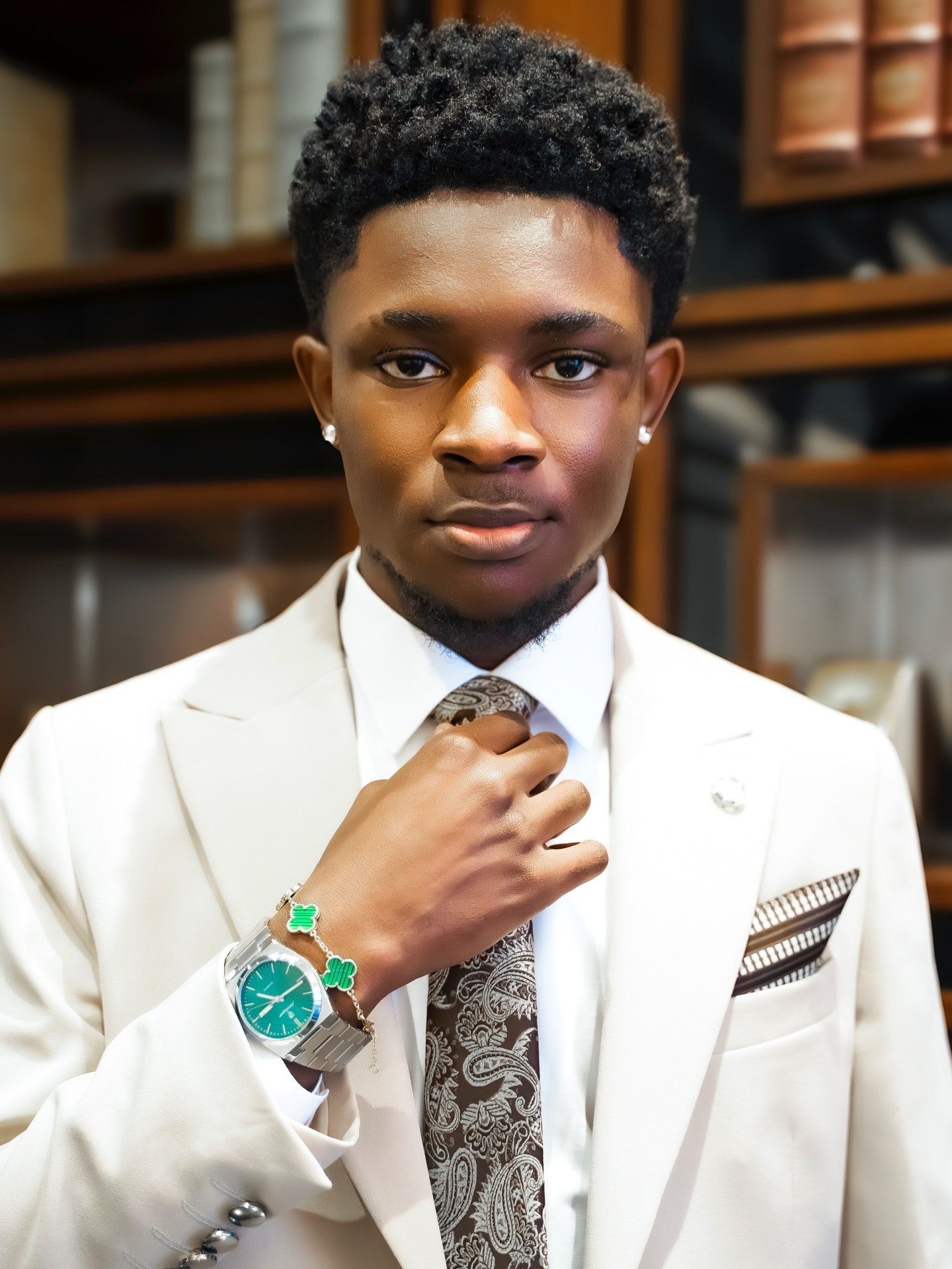 Young man in cream suit adjusts paisley tie while wearing green watch in wood-paneled room.