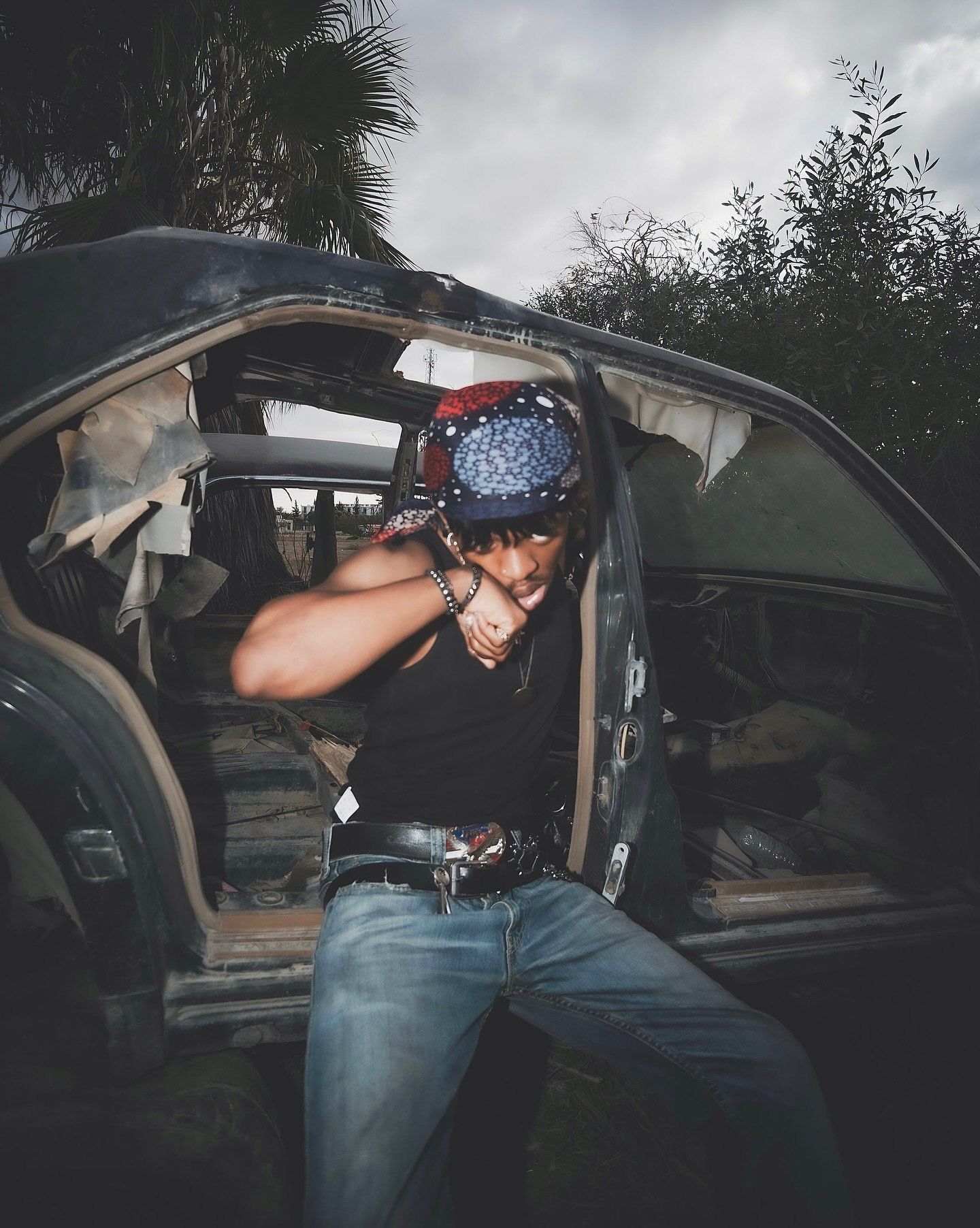 A young man in jeans and bandana poses dramatically beside an abandoned wrecked car outdoors.
