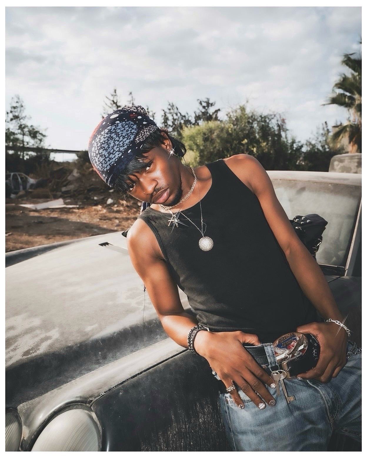 Young man in black tank top and bandana leans on vintage car outdoors, wearing chains and bracelets.