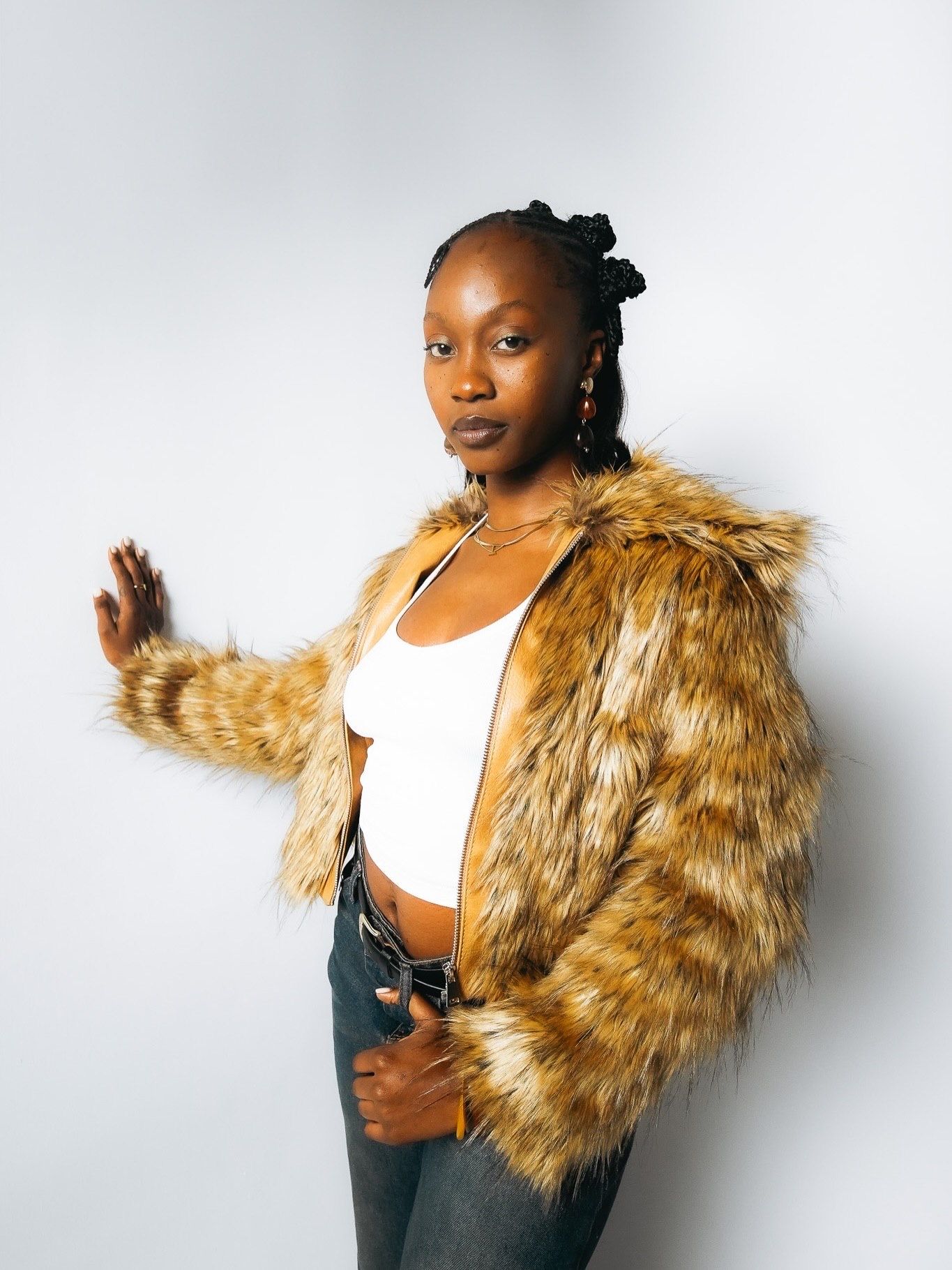 Black woman in golden faux fur coat and white crop top poses confidently against light studio background.