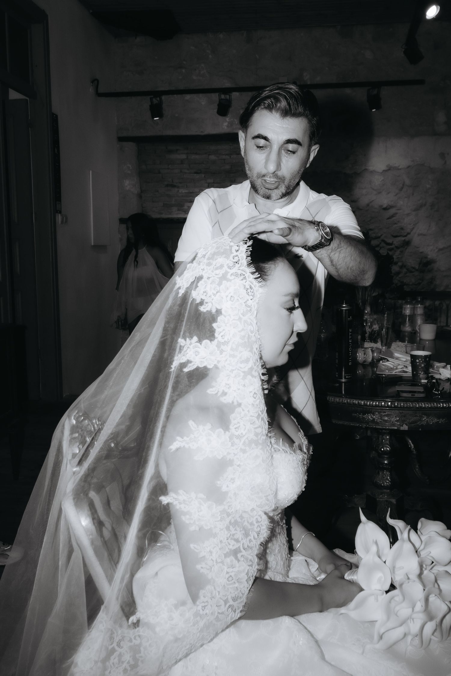 Black and white photo of a groom placing a veil on a bride in a lace wedding dress during bridal preparations.