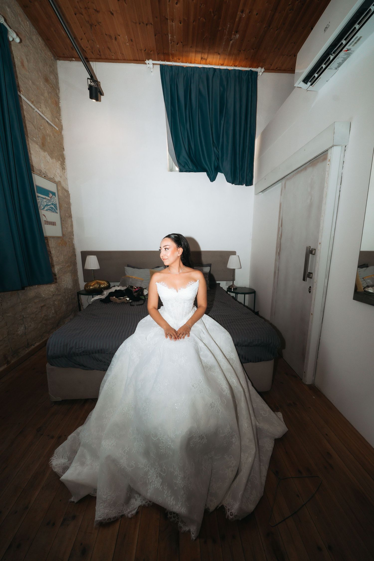 Bride in a white ball gown wedding dress sitting in a rustic bedroom with wooden ceiling and teal curtains.