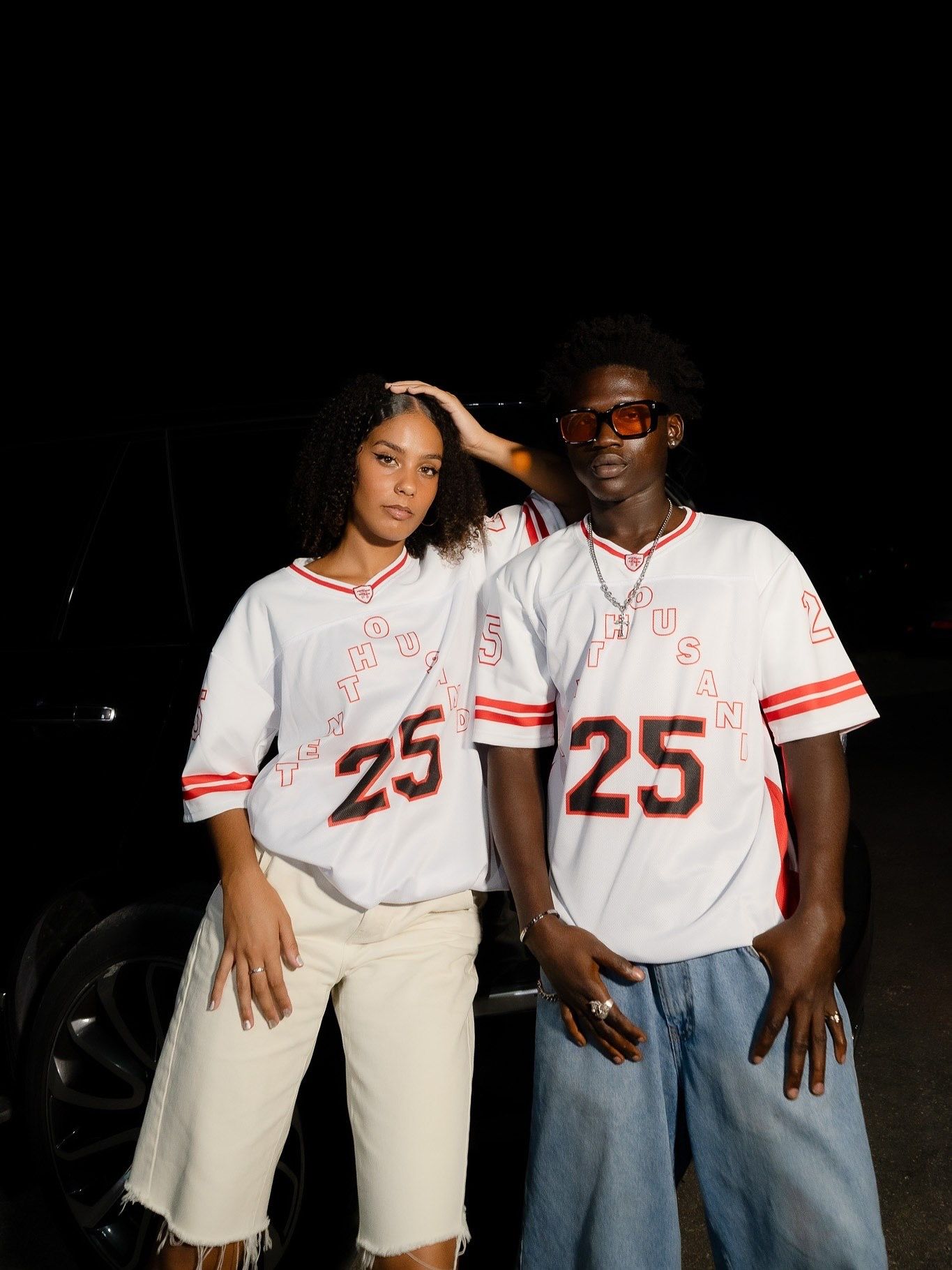 A man and woman wearing matching white and red number 25 Hustle baseball jerseys pose together at night outdoors.