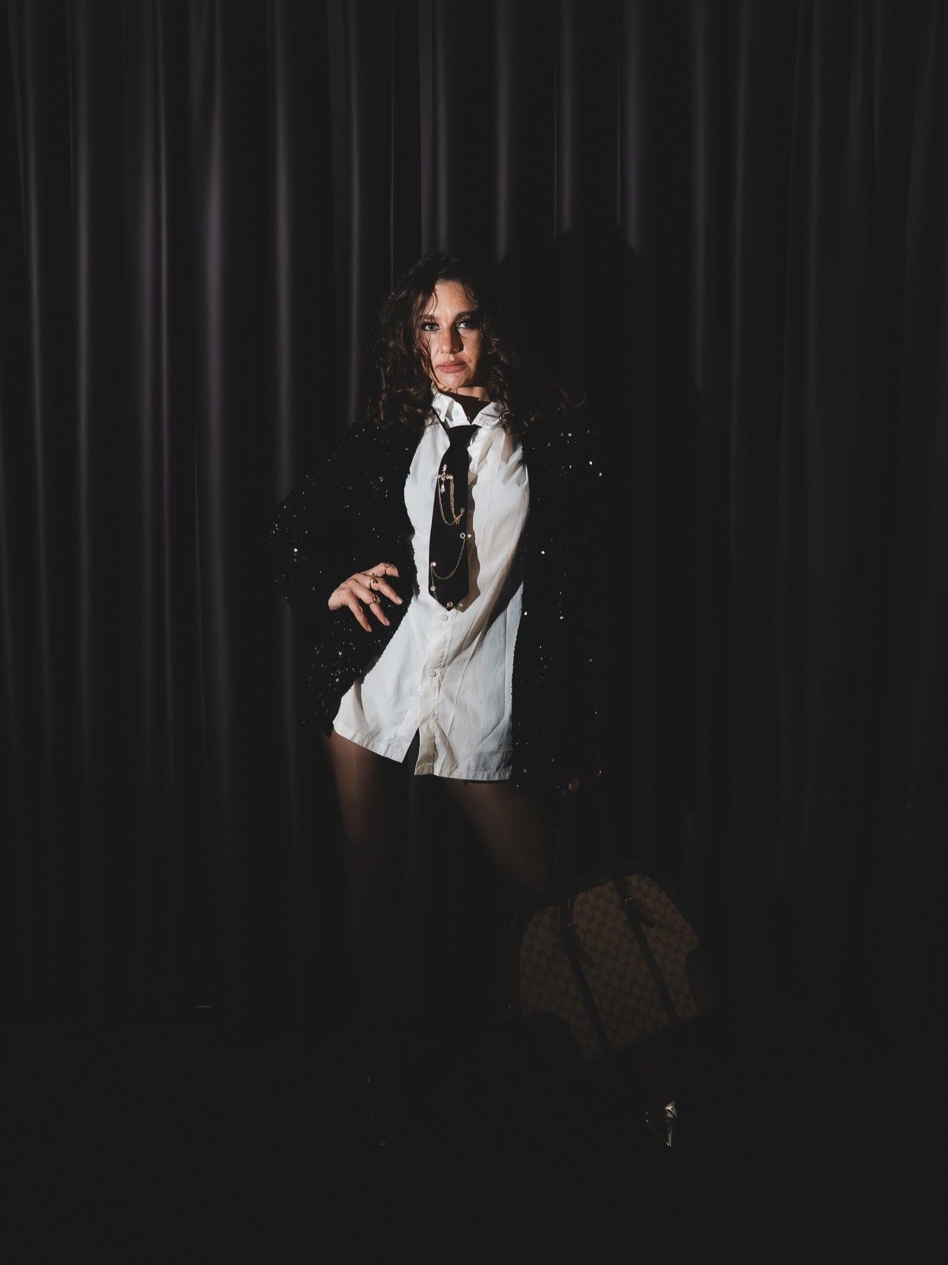 Woman in black blazer white shirt and black tie stands dramatically in dark curtained studio.