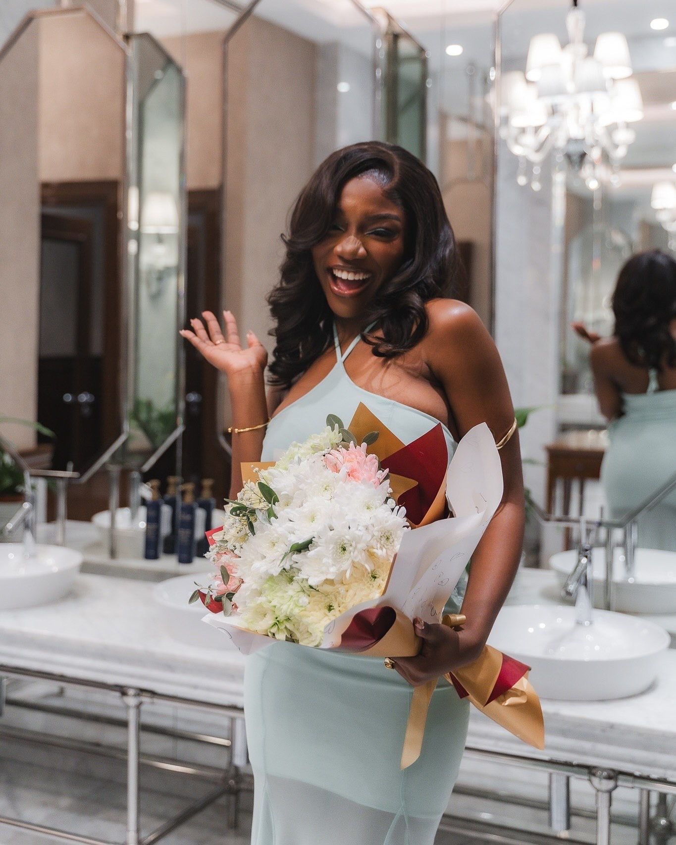 Smiling woman in elegant dress holding white flower bouquet, waving in luxurious marble bathroom setting.