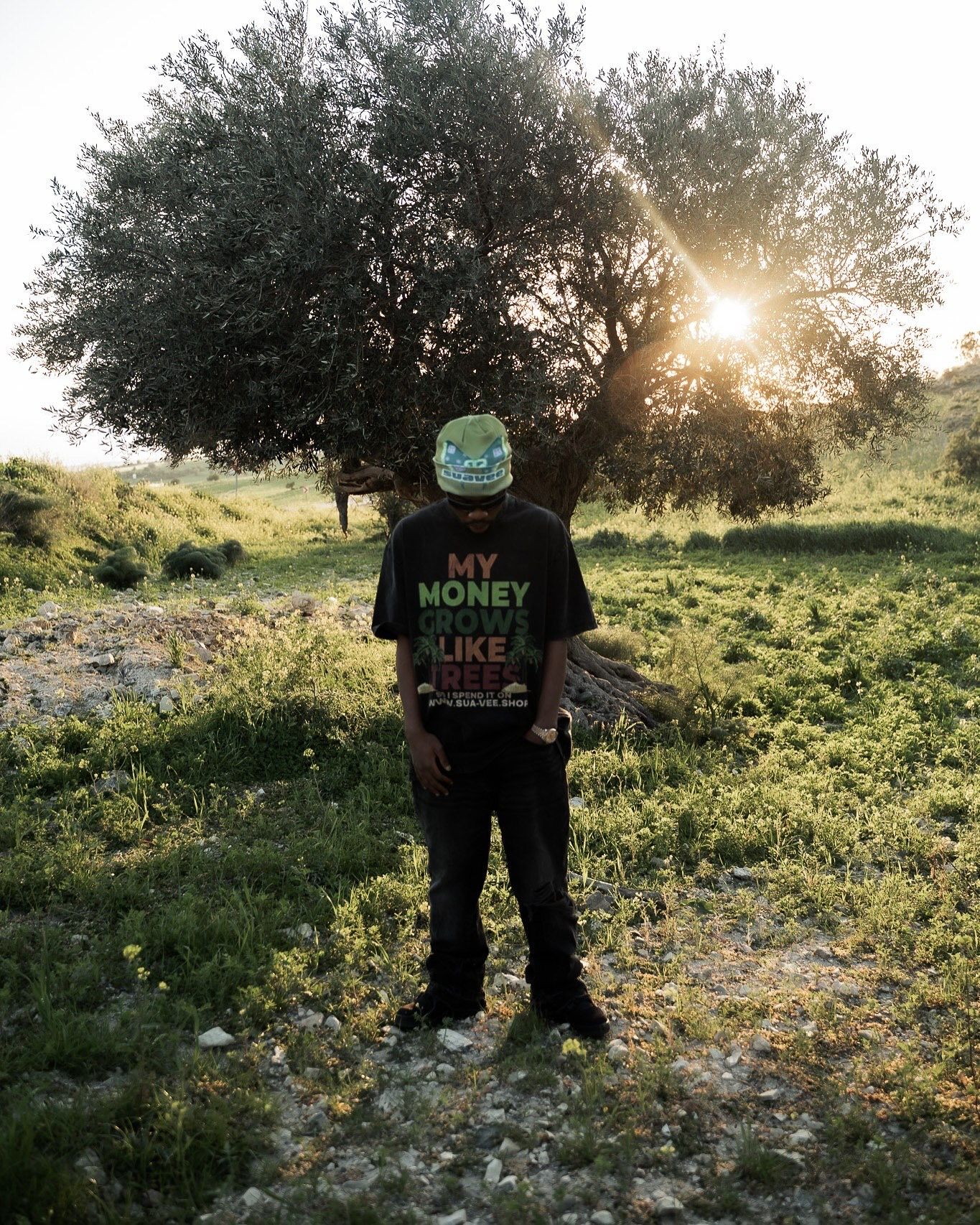 Man standing outdoors in a black graphic tee and green cap, backlit by golden sunset light near a large tree.