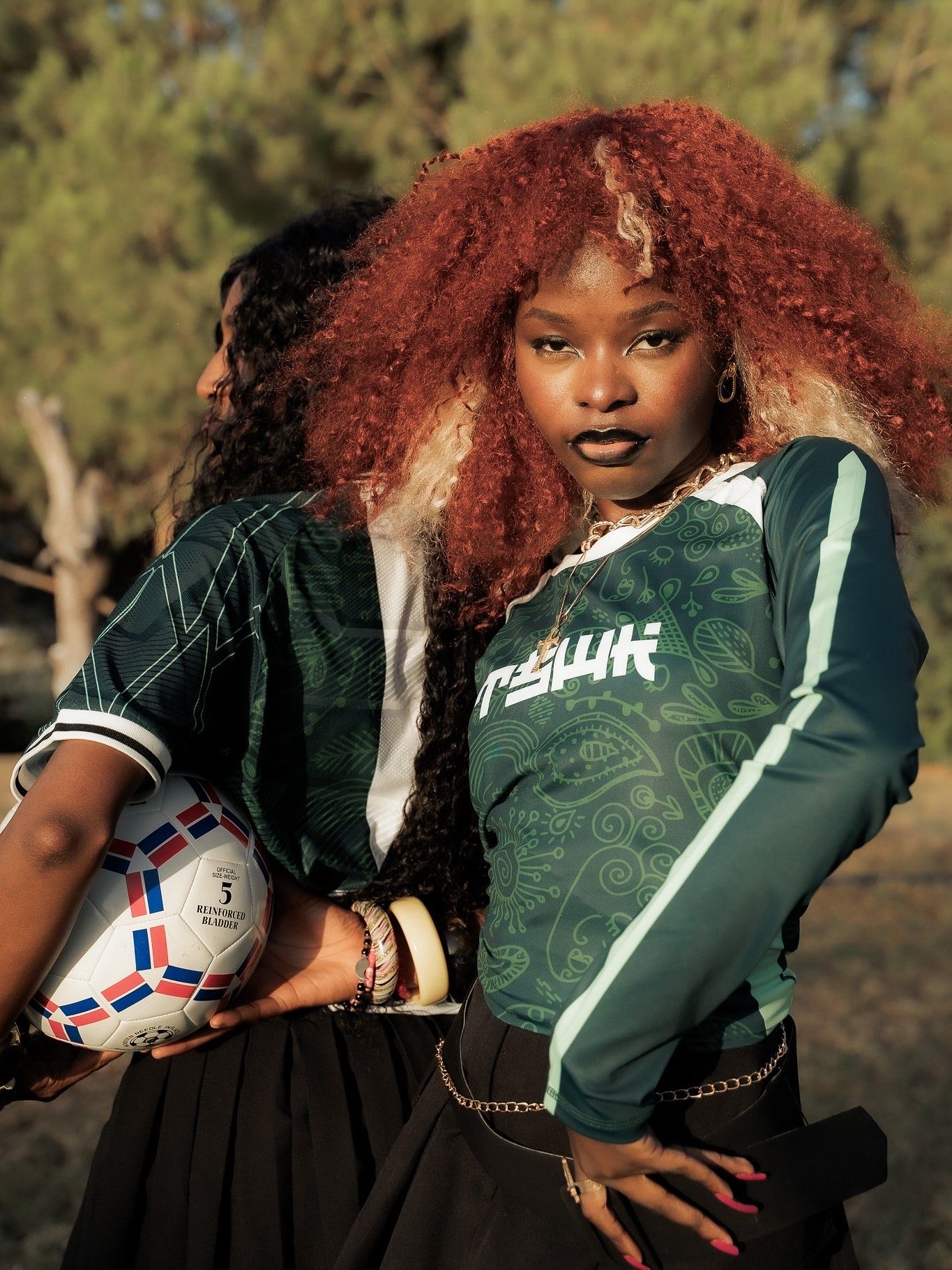 Two young women in green soccer jerseys, one with red curly hair holding a soccer ball, posing outdoors.