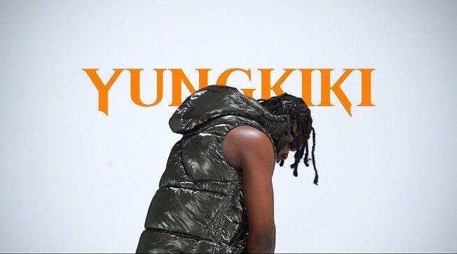 Yungkiki artist in camo puffer vest with dreadlocks, head bowed, against white background with orange text.