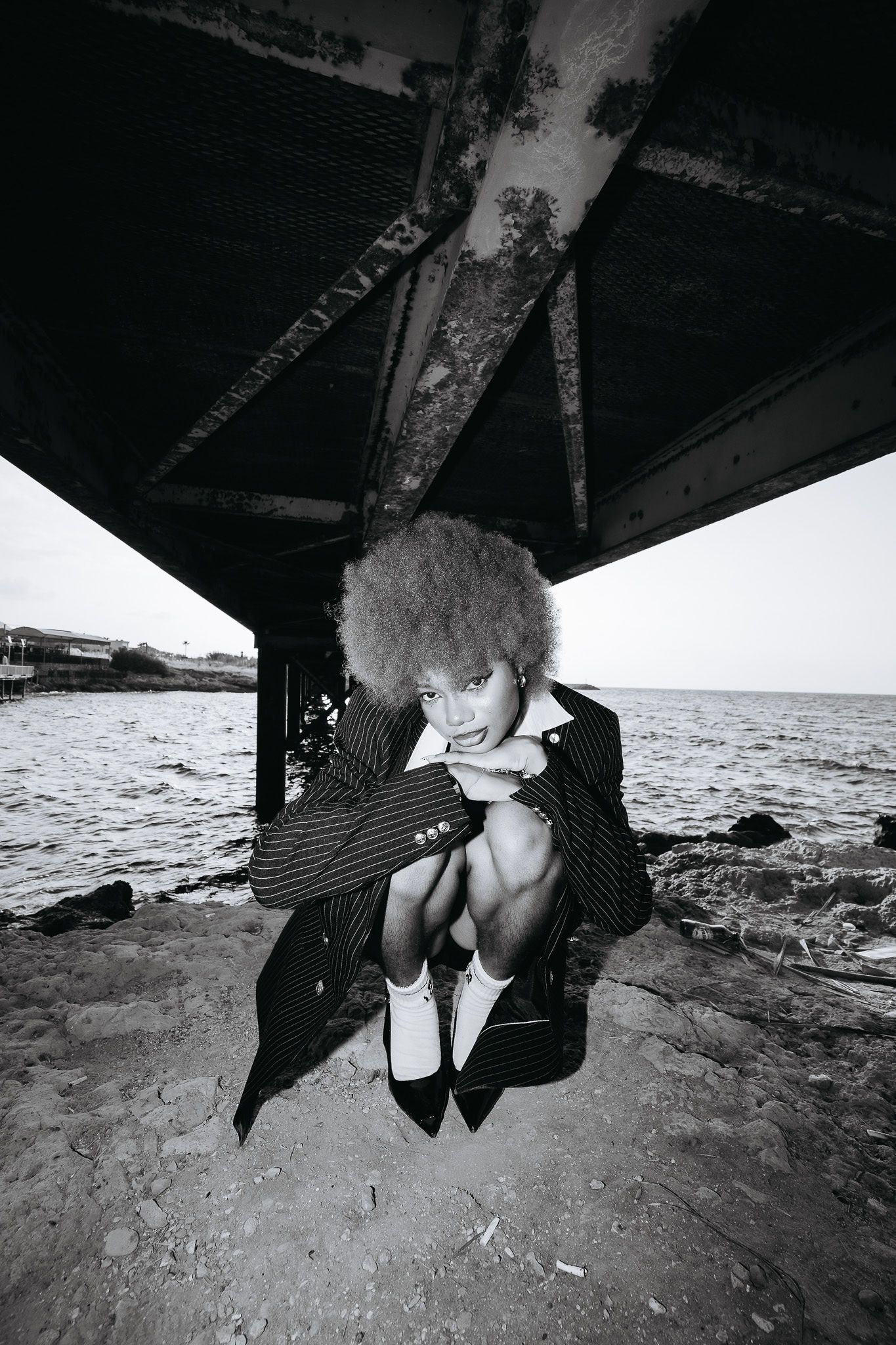Black and white photo of a woman in a pinstripe suit crouching under a rusty pier by the ocean.