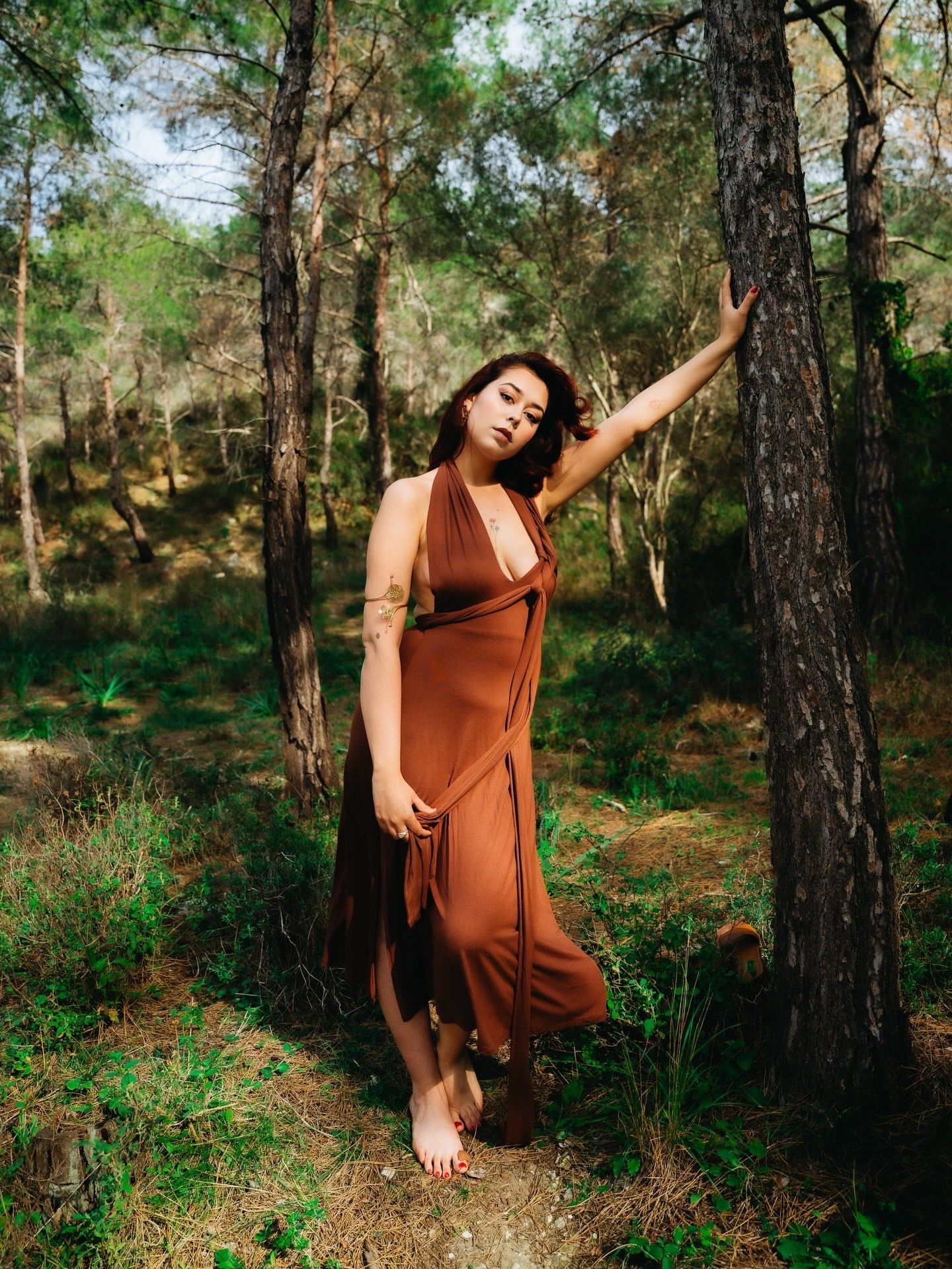 Woman in a rust-colored halter maxi dress posing barefoot in a sunlit pine forest.