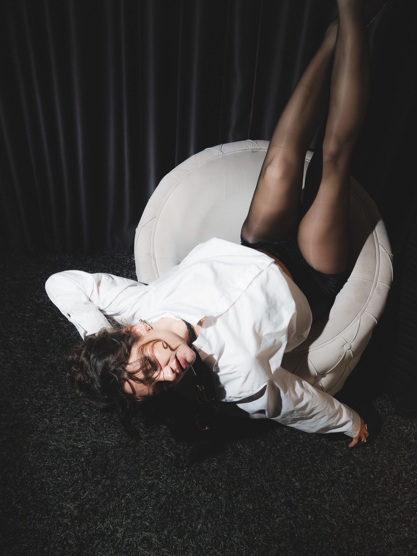 Woman in white shirt and black tights reclines against white chair on dark floor in artistic pose.