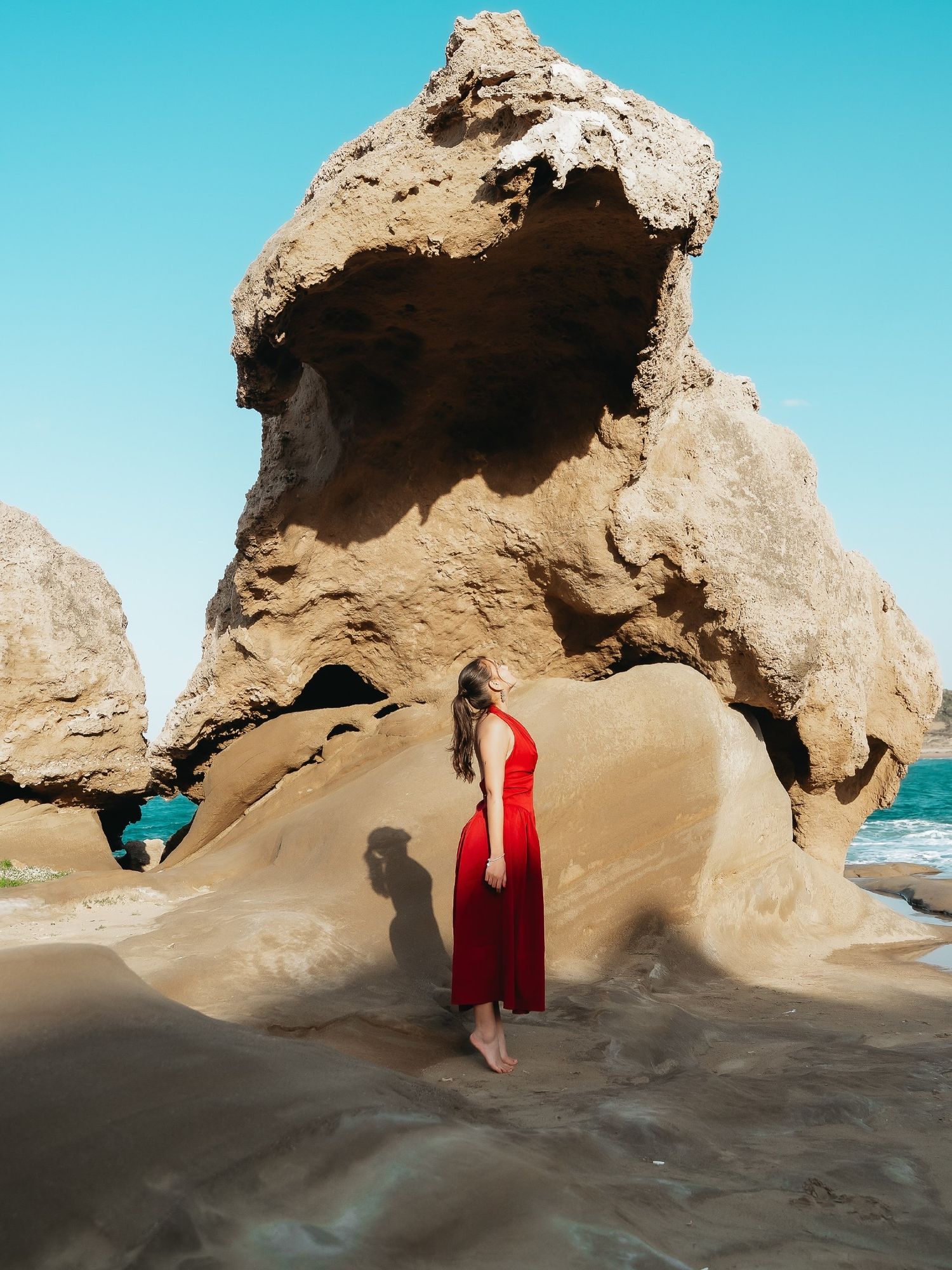 Woman in red dress standing among large sandstone rock formations on a sunny beach with turquoise water.