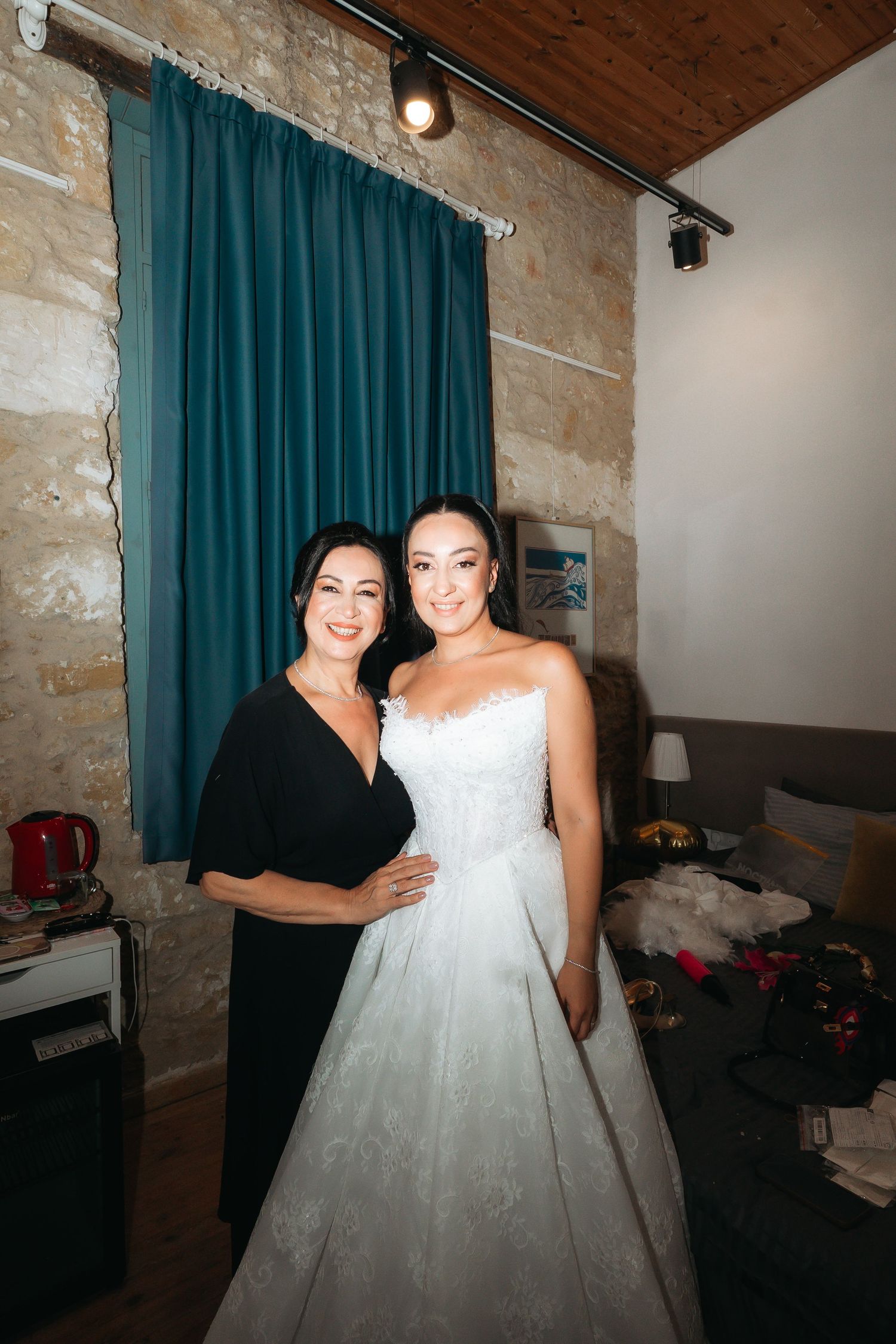 Bride in white lace wedding gown poses with woman in black dress in rustic stone-walled room with teal curtains.