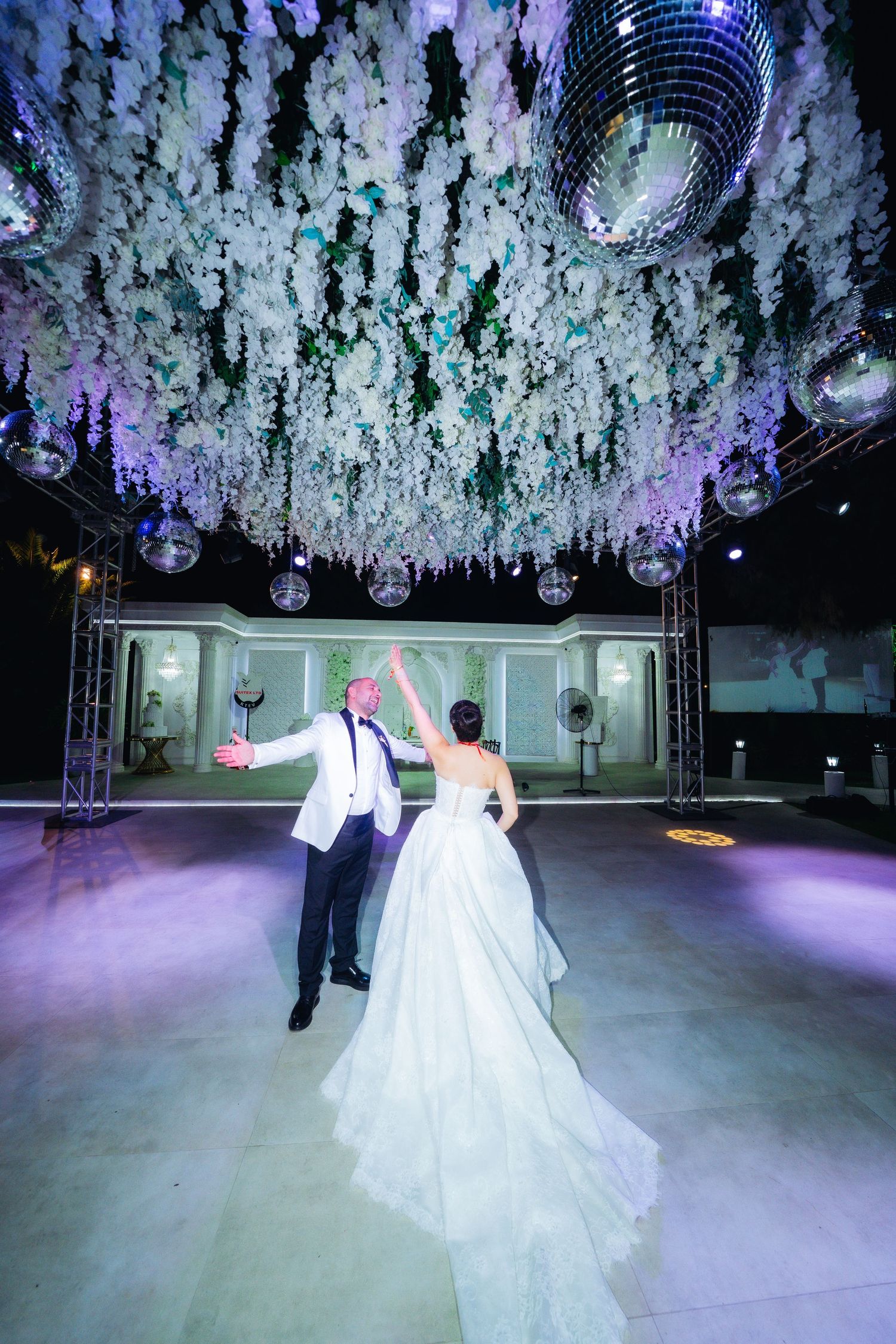 Bride and groom share first dance under floral ceiling with disco balls at elegant outdoor wedding reception.