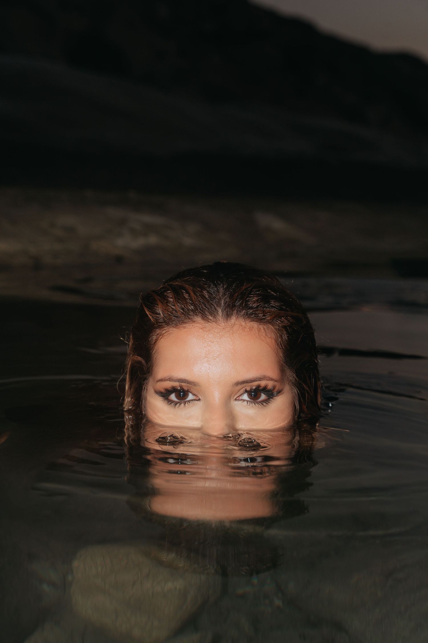Woman with wet hair and dramatic eye makeup submerged in dark water, eyes visible above the surface.