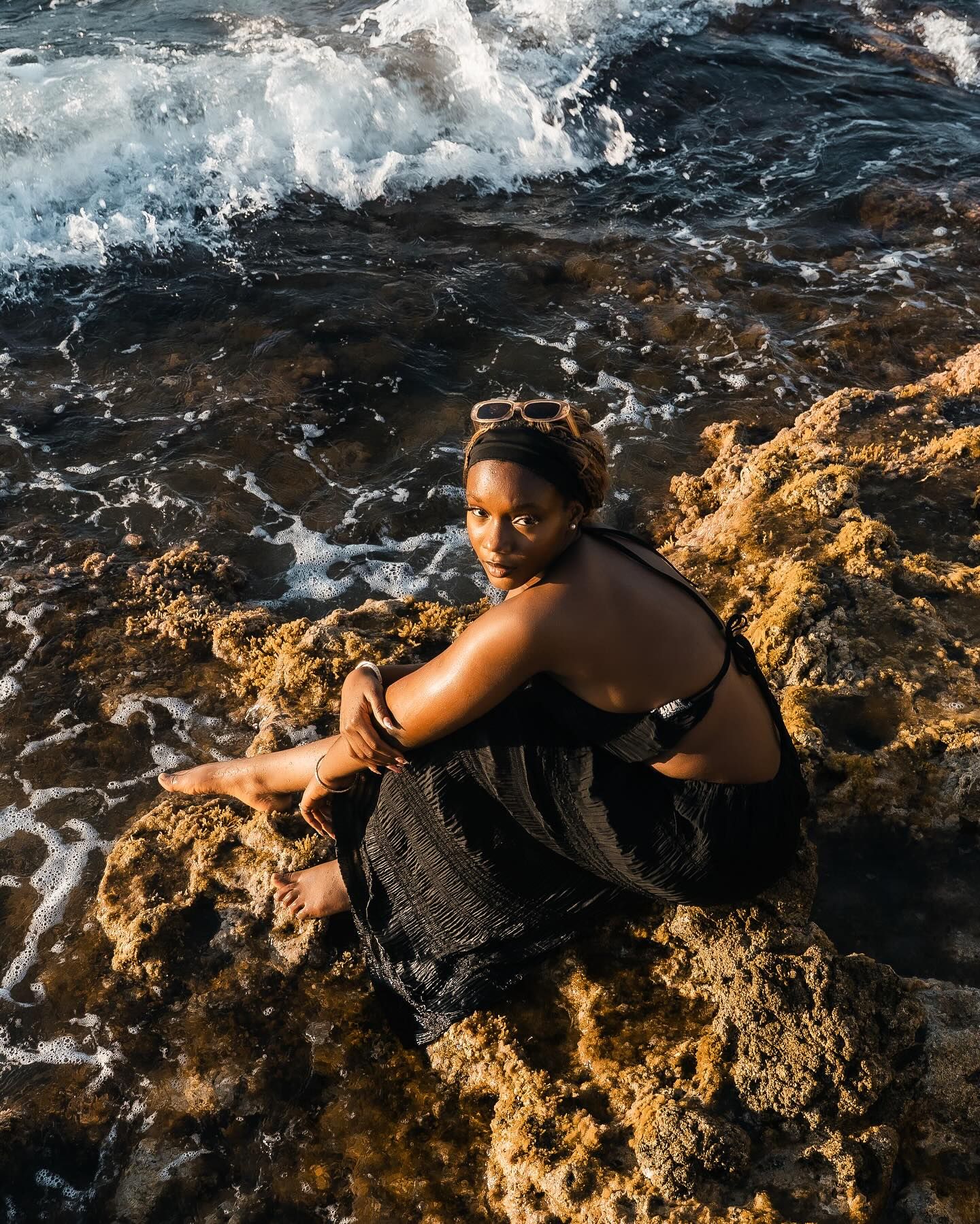 Woman in black dress sitting on rocky shoreline with ocean waves crashing behind her.