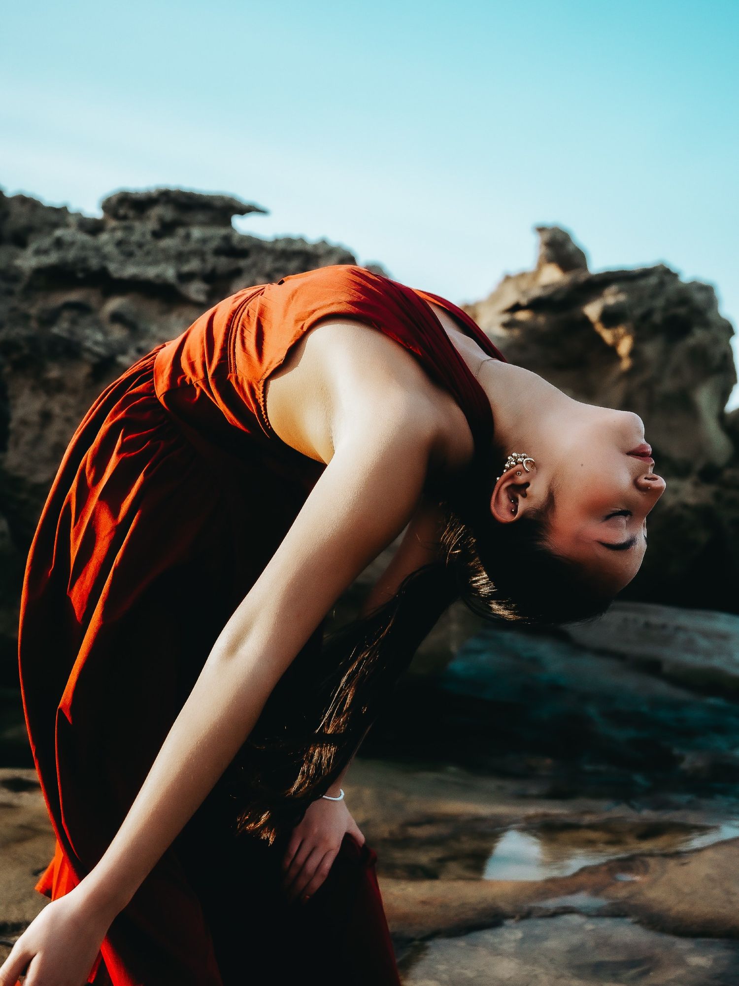 Woman in rust-red dress leans back dramatically on rocky coastal terrain under a blue sky.