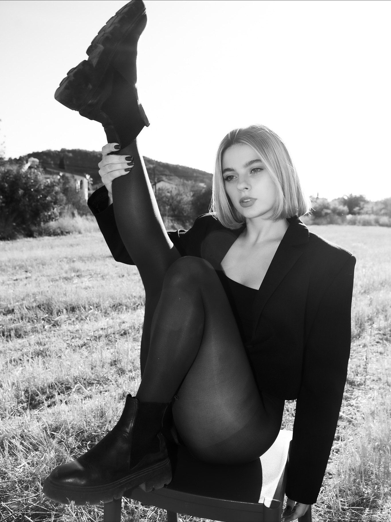 Black and white photo of a blonde woman in a blazer and tights posing on a chair in a field.