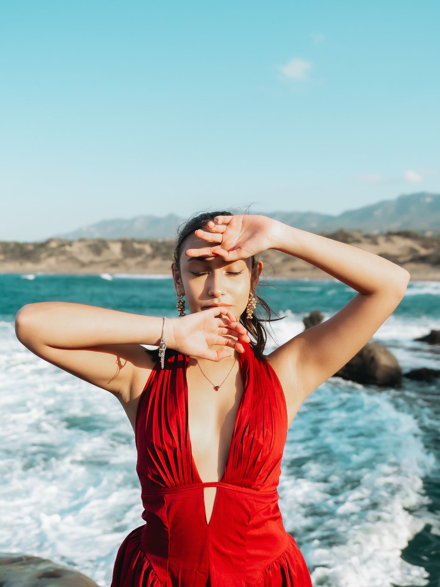 Woman in red halter dress posing with hands framing face near rocky ocean shore with mountains in background.