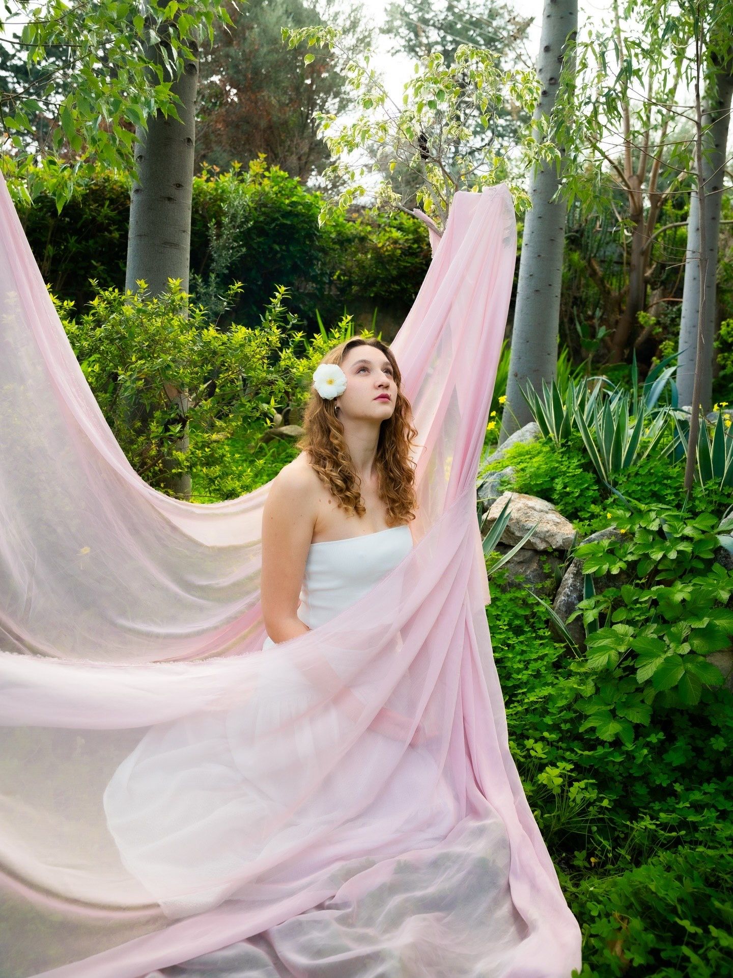 A bride in a white strapless gown with a floral headpiece poses outdoors, surrounded by lush greenery and flowing pink fabric.