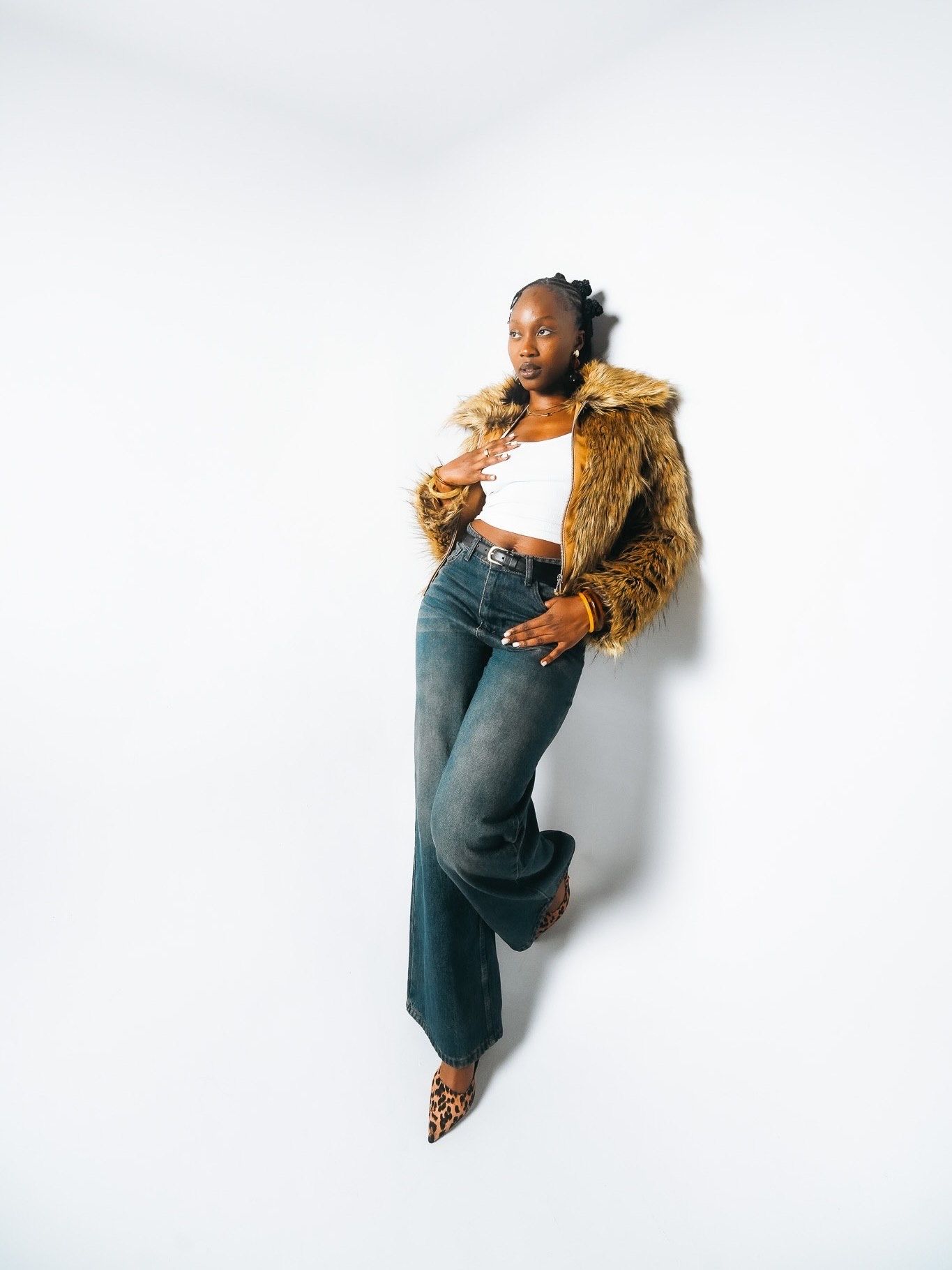 Black woman in faux fur coat, white crop top and wide-leg jeans poses against white studio background.