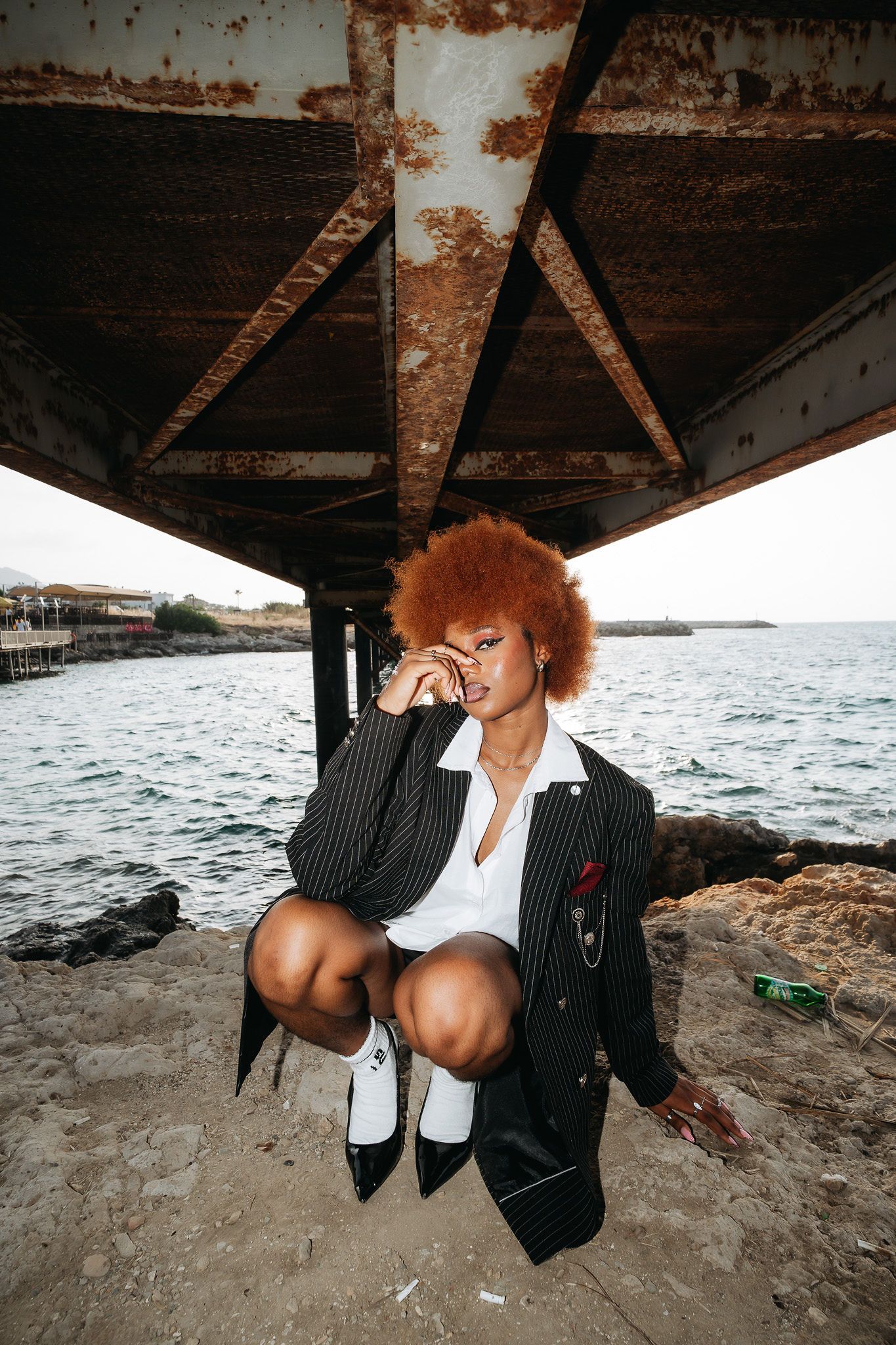Woman with auburn afro in pinstripe suit crouches under rusty pier structure by the sea.