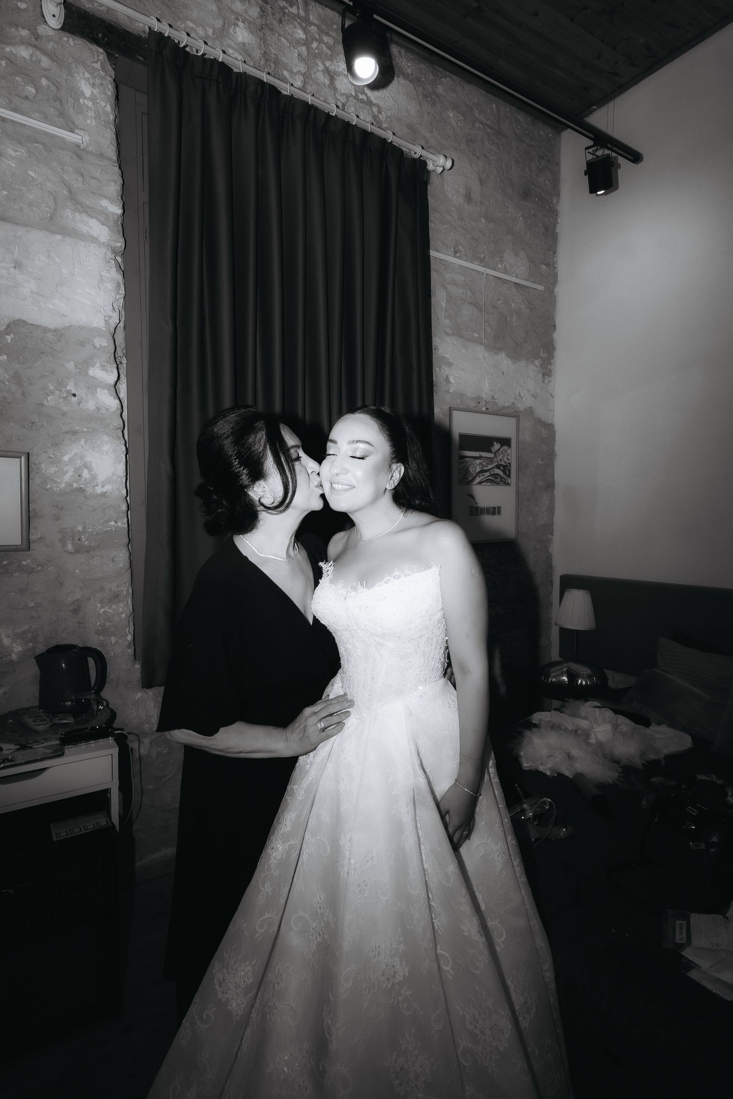 Black and white photo of bride in white gown receiving a kiss on cheek from woman in black dress indoors.