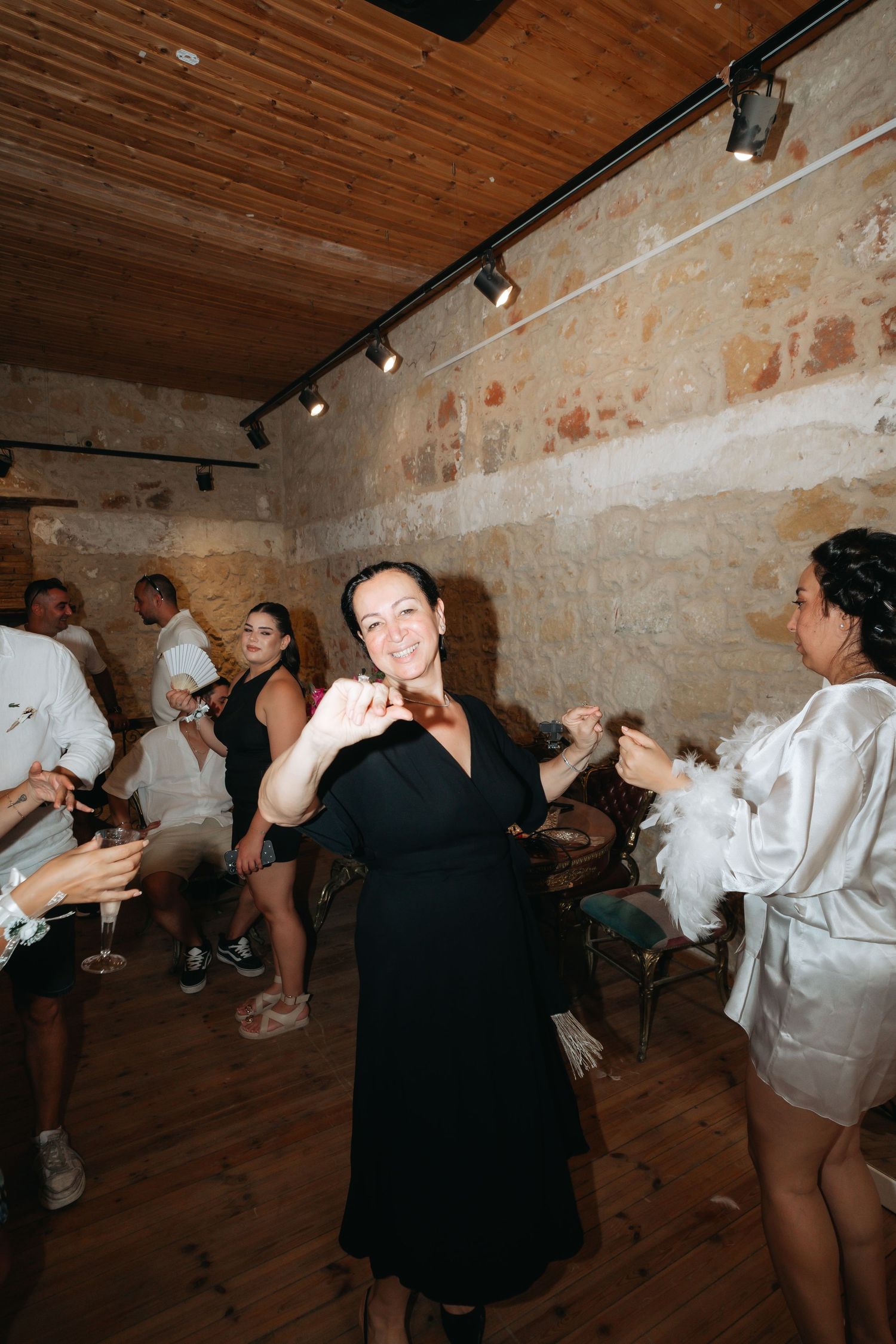 Woman in black dress dancing joyfully at a rustic wedding reception with stone walls and wooden ceiling.