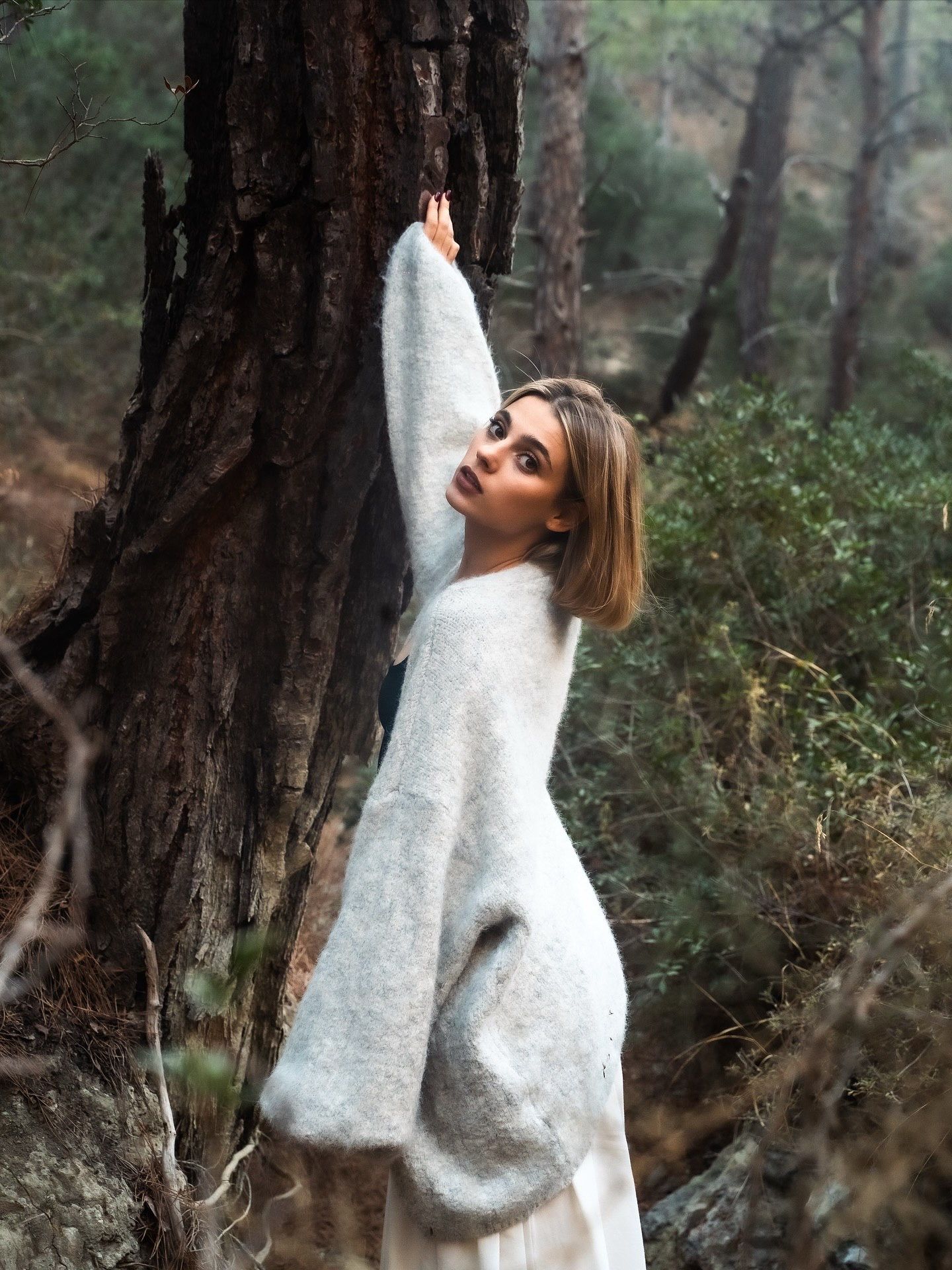 Woman in white fuzzy sweater leaning against a tree trunk in a misty forest setting.