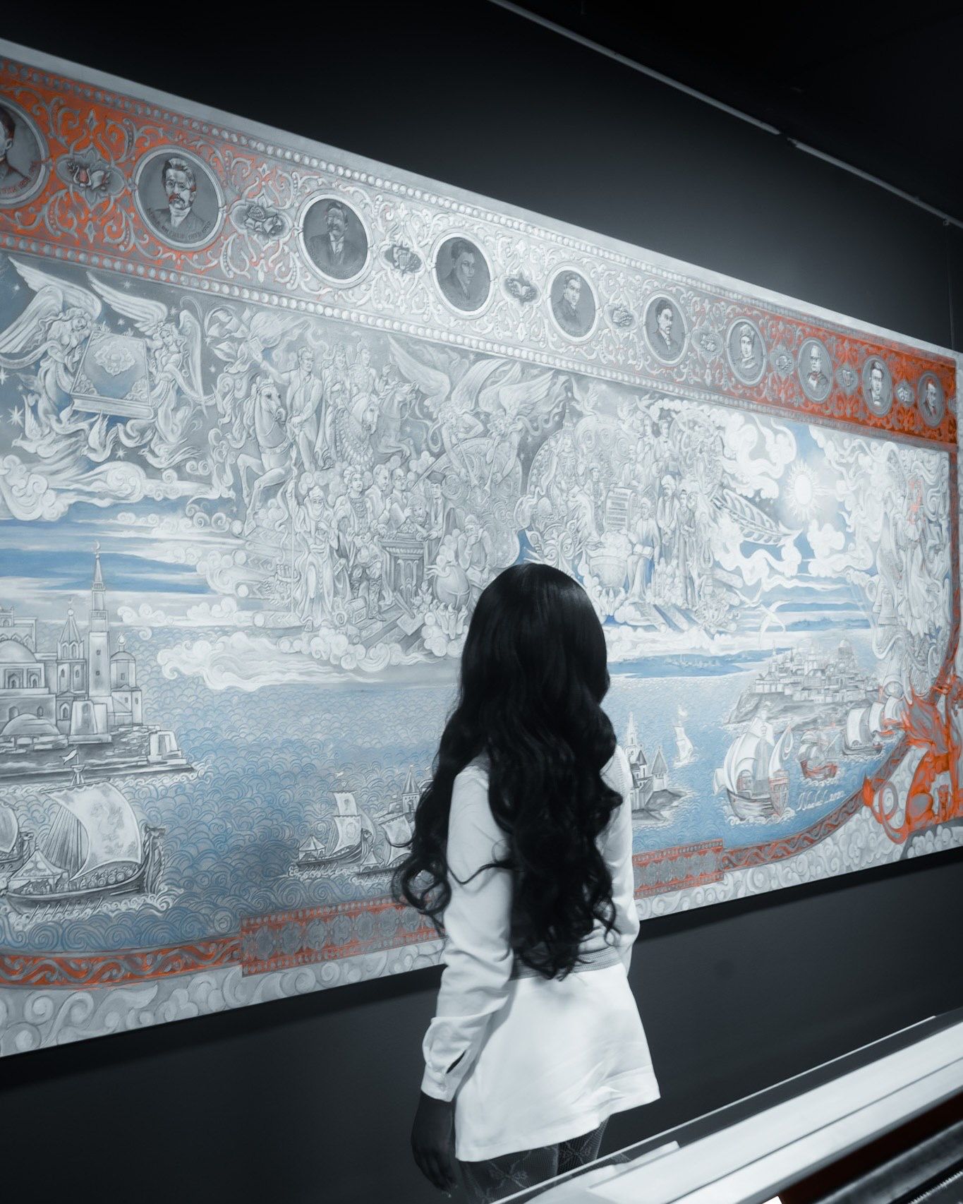 Woman with long black hair views an ornate tapestry artwork featuring ships and mythological scenes in a museum.