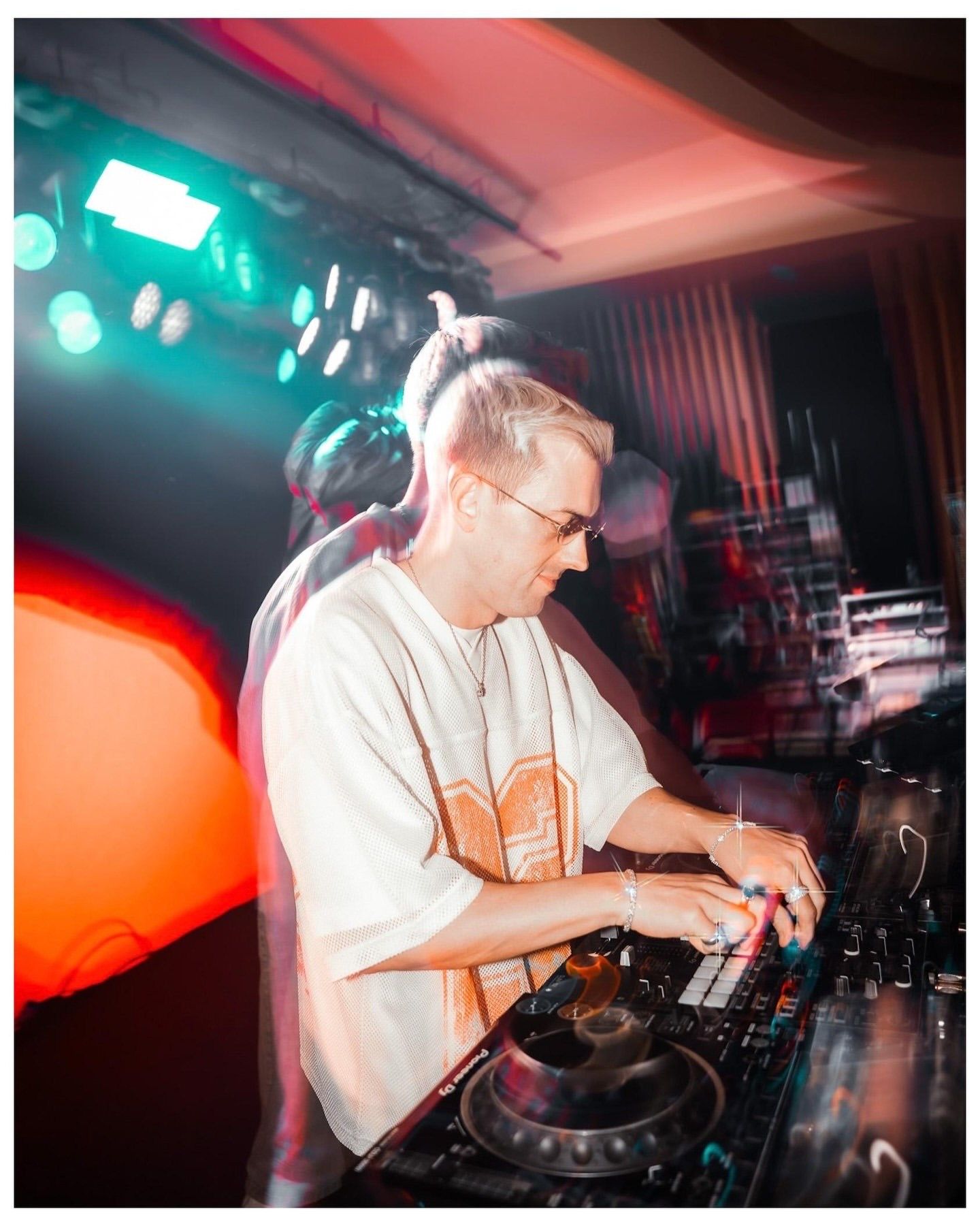 DJ with blonde hair and glasses mixing at turntables under colorful stage lights in a nightclub.