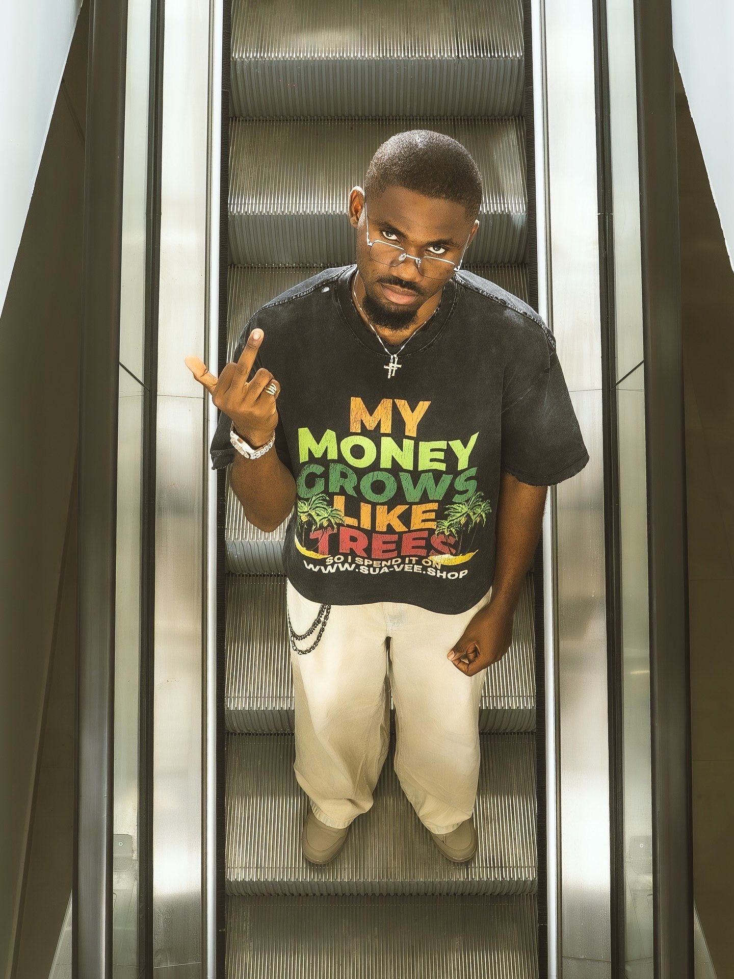 Man on escalator wearing black graphic tee reading My Money Grows Like Trees, beige pants, making hand gesture.