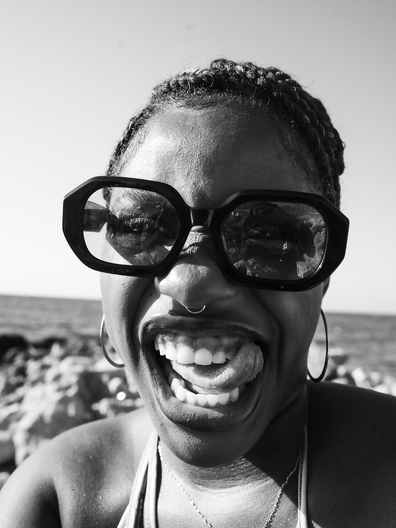 Black and white photo of a smiling woman wearing large sunglasses and hoop earrings at a beach.