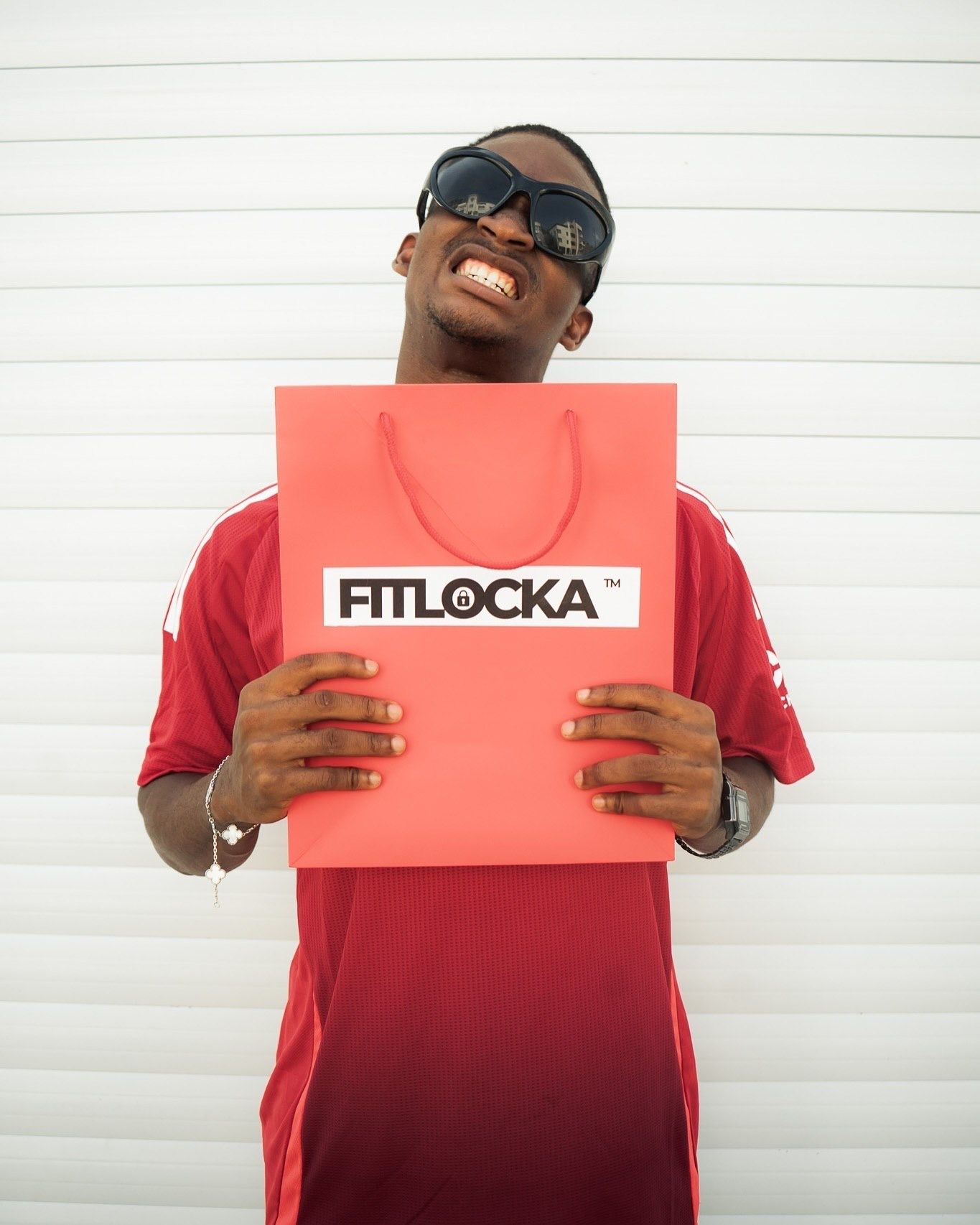 Man in red jersey and sunglasses holding a coral FITLOCKA branded shopping bag.