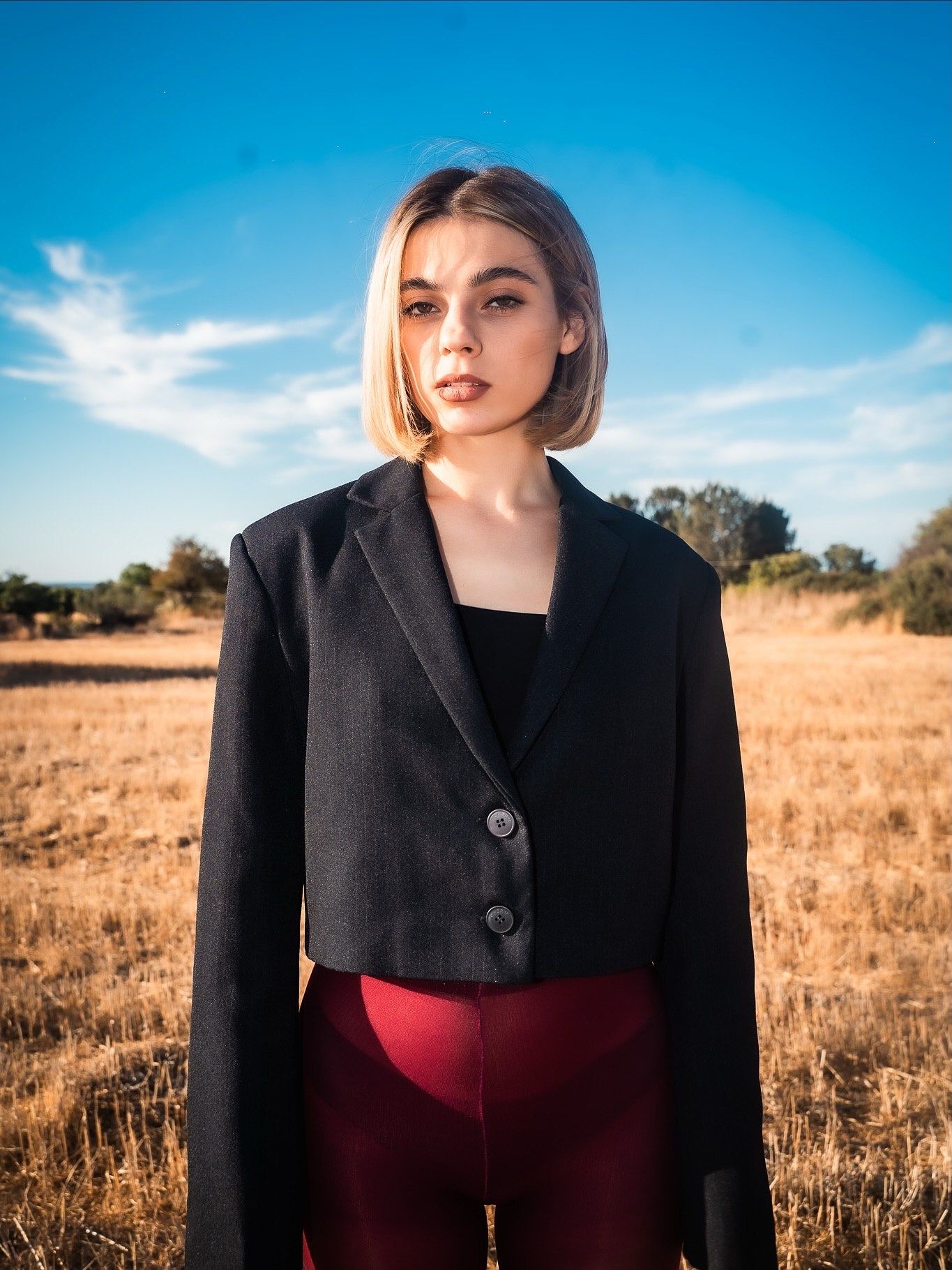 Young woman in cropped black blazer and burgundy pants standing in golden field under blue sky.