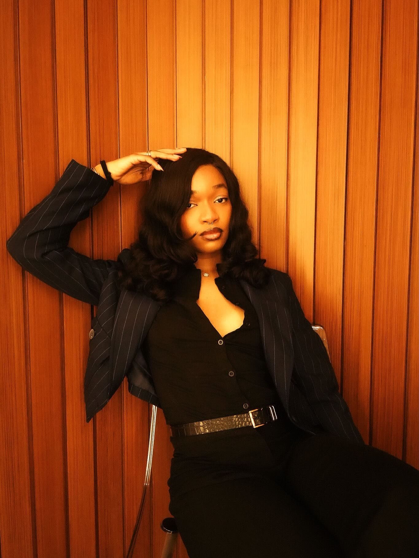 Young woman in black outfit and blazer poses confidently against warm wood-paneled wall.