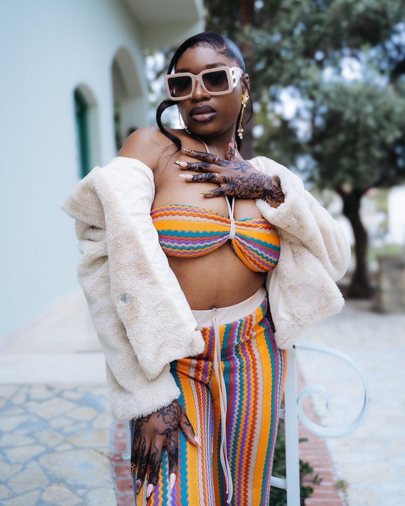Woman in colorful striped crop top and pants with fur coat and sunglasses, henna tattoos on hands, outdoor setting.