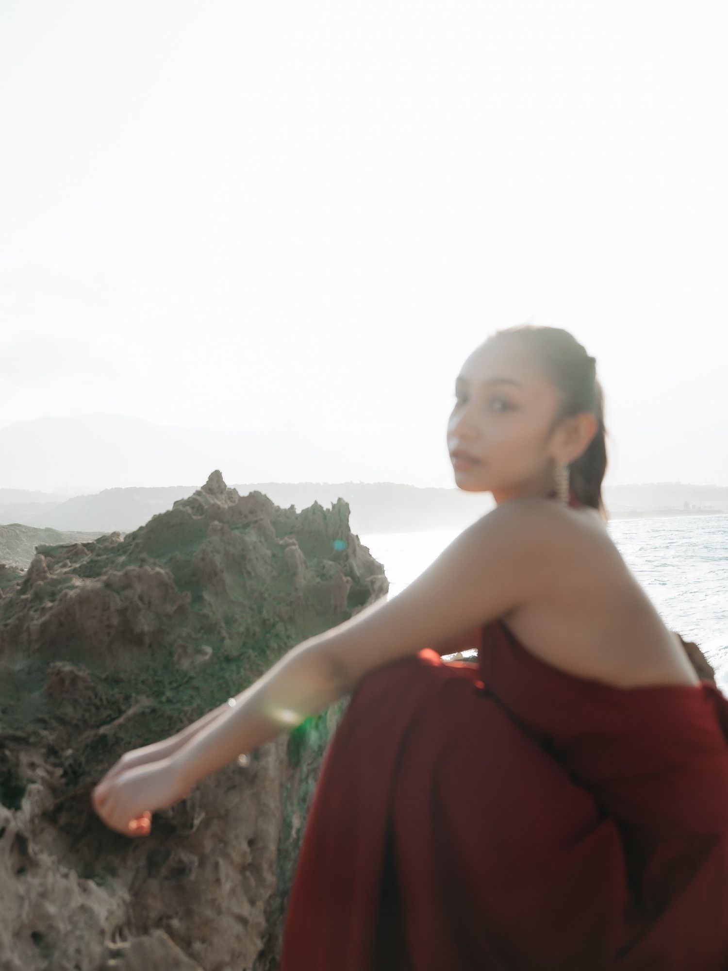 Woman in red dress sitting on rocky coastal cliff with ocean and misty mountains in background.