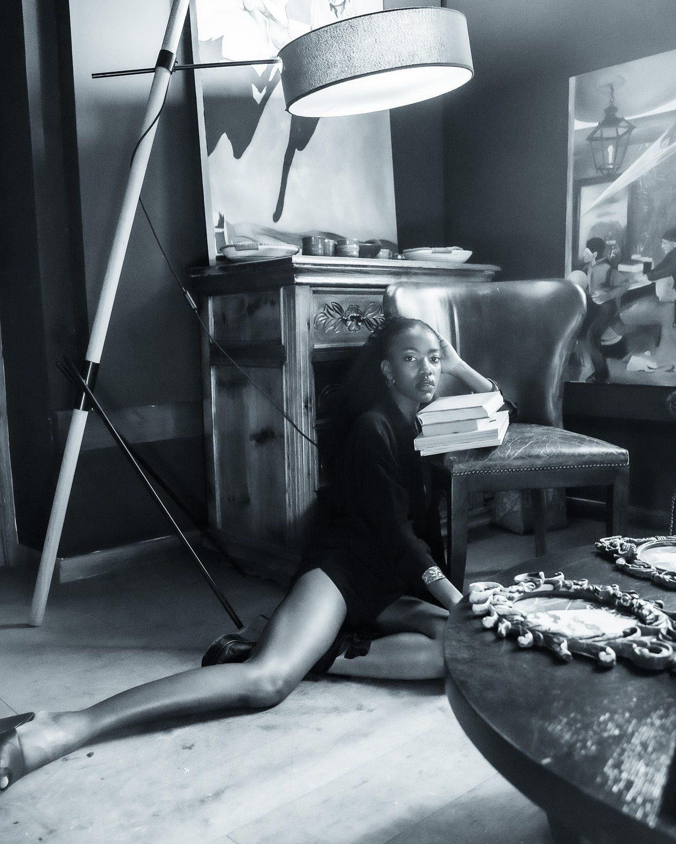 Black and white photo of a woman sitting on the floor leaning against books stacked on a chair in an elegant room.