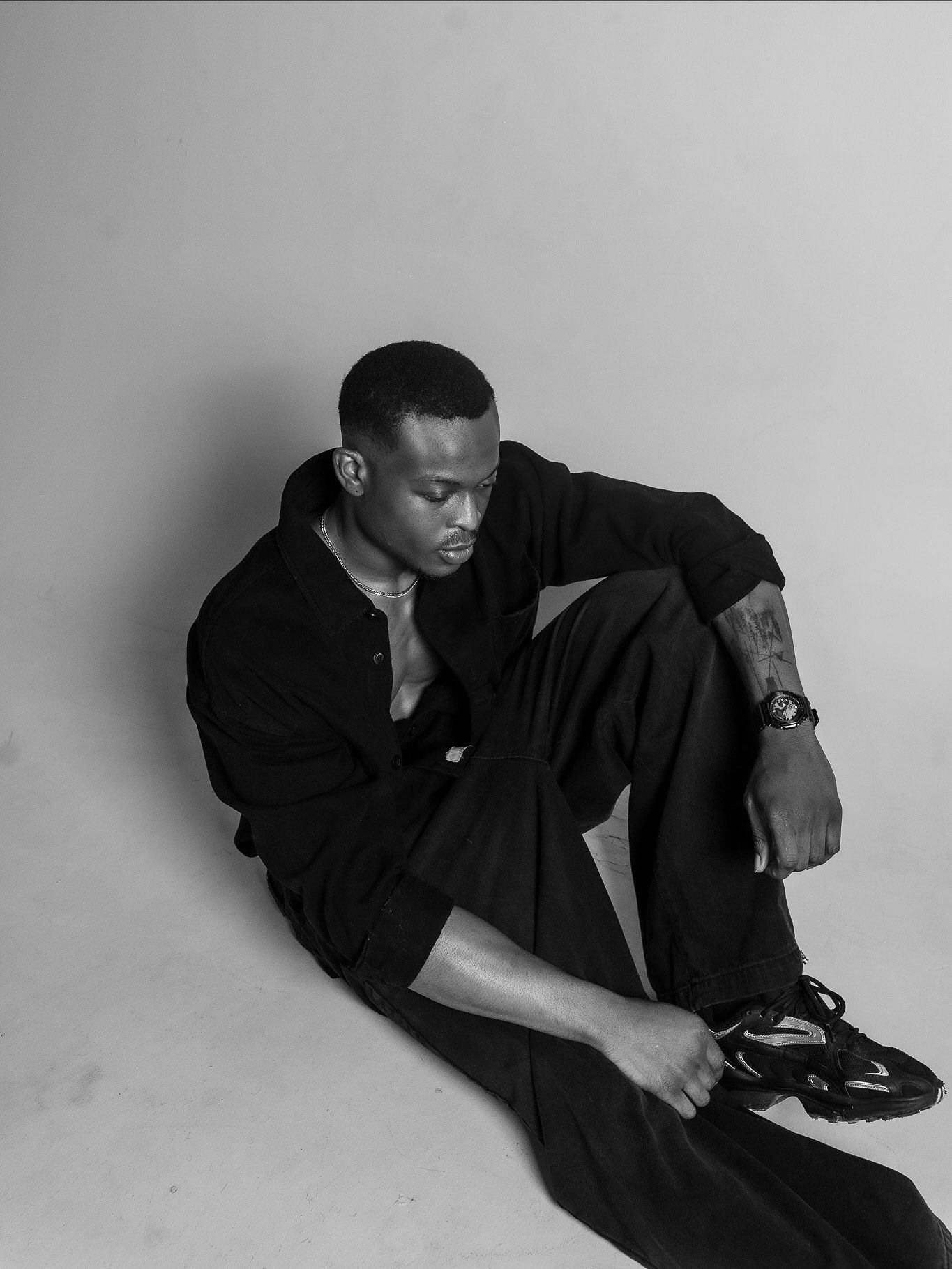 Black and white photo of a young man in all-black outfit sitting on floor, looking down thoughtfully.