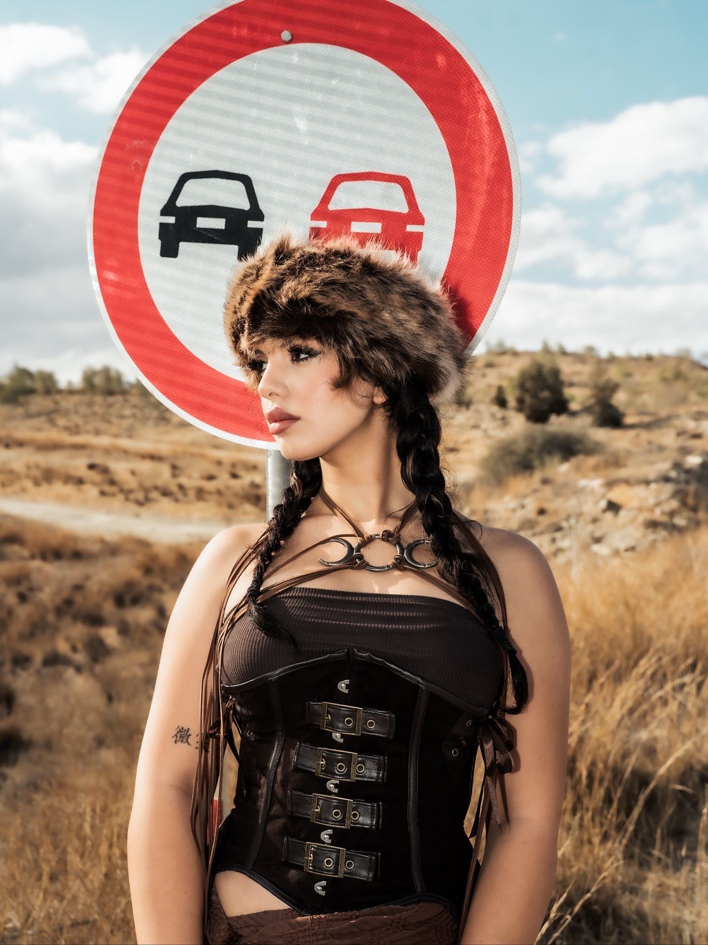 Woman in fur hat and black corset poses before a no-overtaking road sign in a arid landscape.