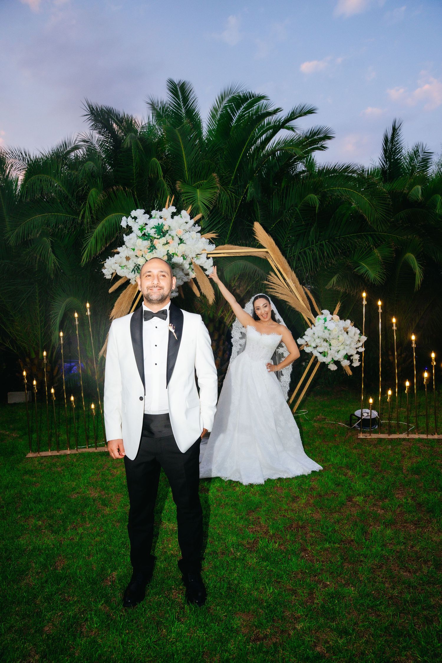Bride and groom pose outdoors at tropical wedding ceremony with floral arch, palm trees, and candlelit aisle at dusk.