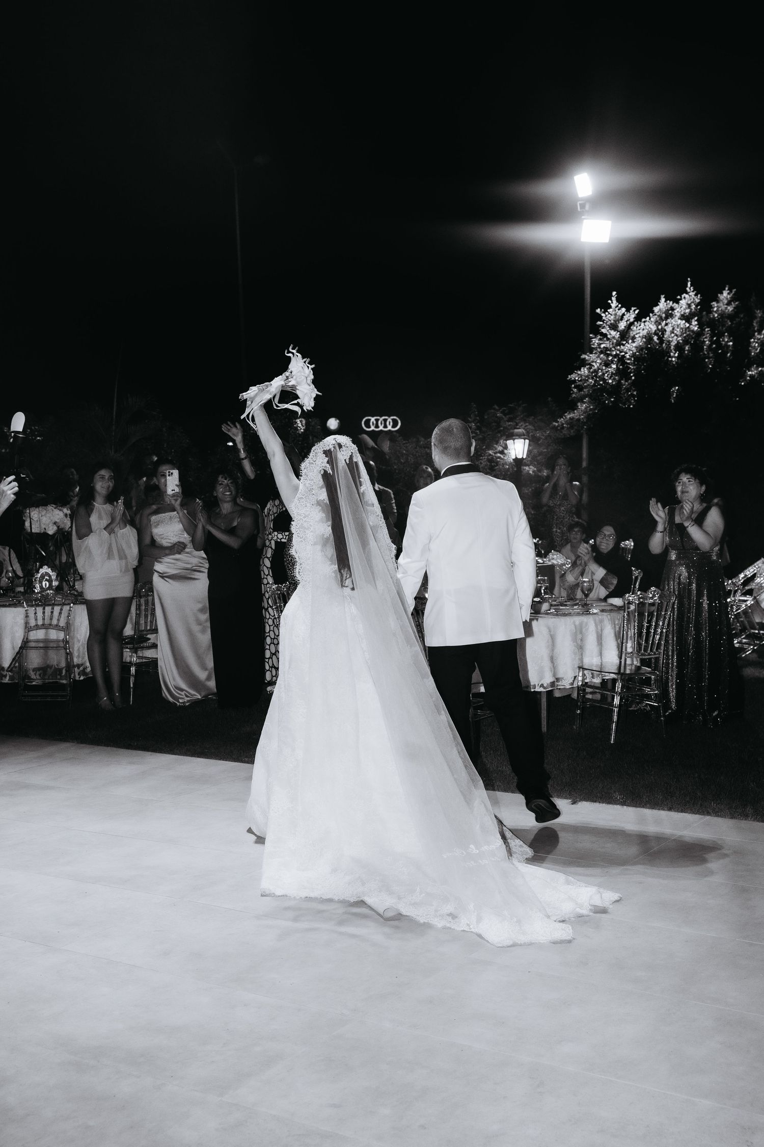 Black and white photo of bride raising bouquet while walking with groom on dance floor at outdoor wedding reception.
