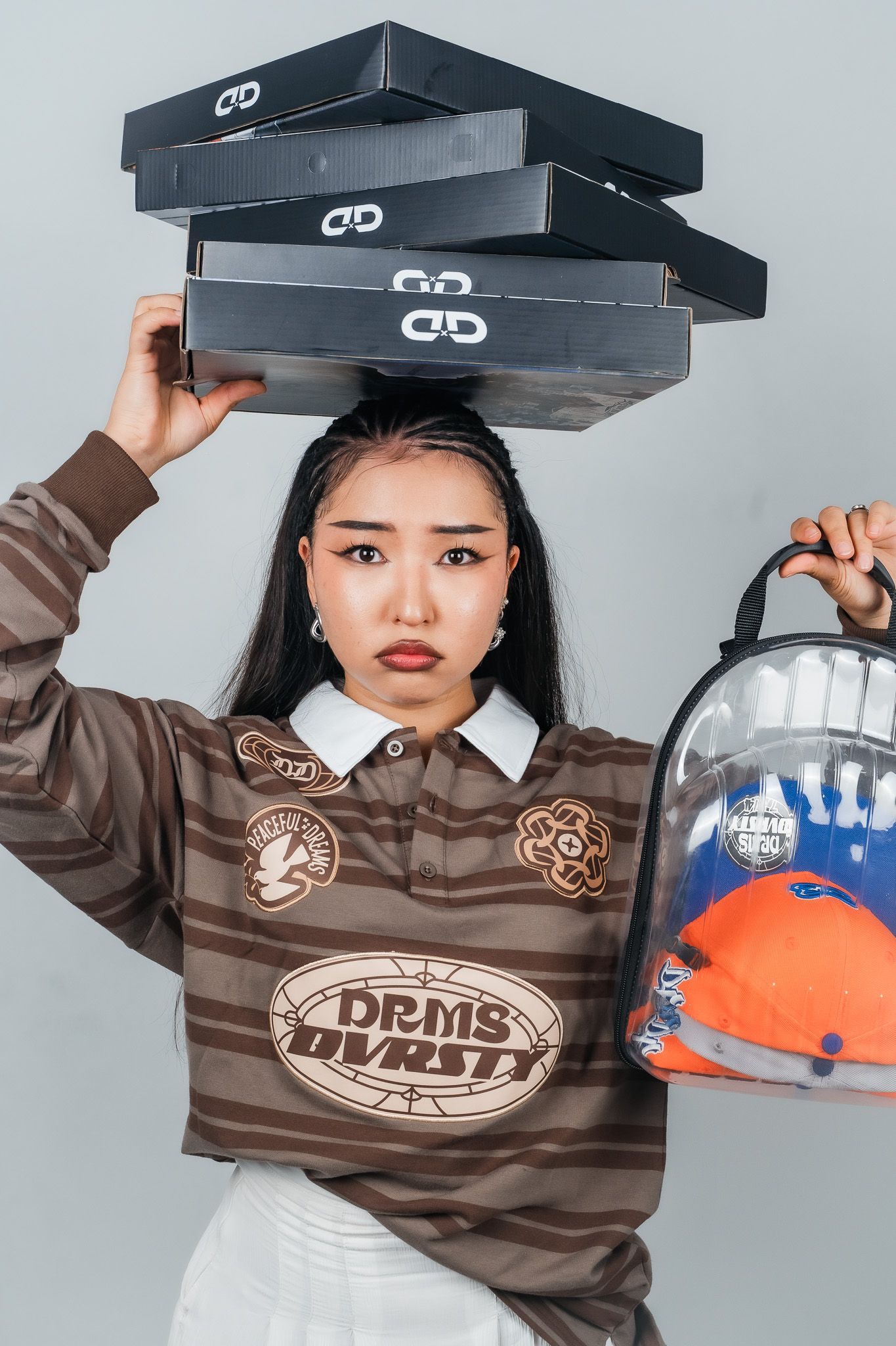 Young woman balancing shoe boxes on head, holding clear backpack, wearing DRMS VARSITY striped brown jersey.